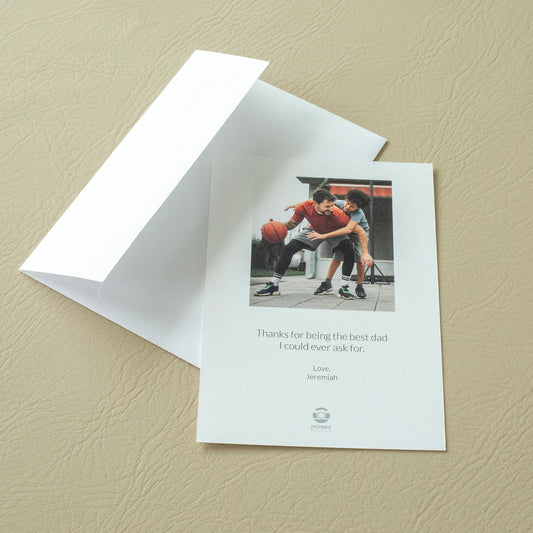 Custom flat Father's Day card and white envelope on a leather surface, featuring a photo of a father and teenage son playing basketball together with the personalized message 'Thanks for being the best dad I could ever ask for'