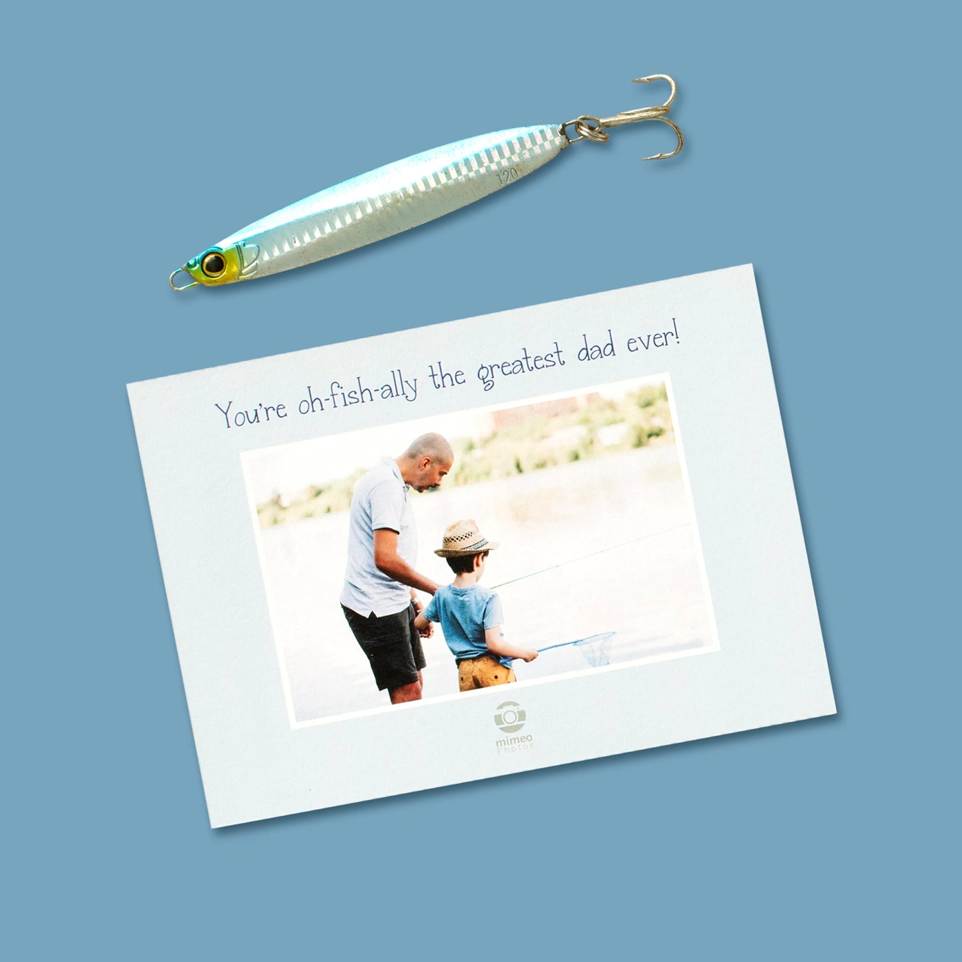 Custom flat Father's Day card on a blue surface beside a fishing lure, reading 'You're oh-fish-ally the greatest dad ever!' with a photo of a father teaching his young son to fish by the water