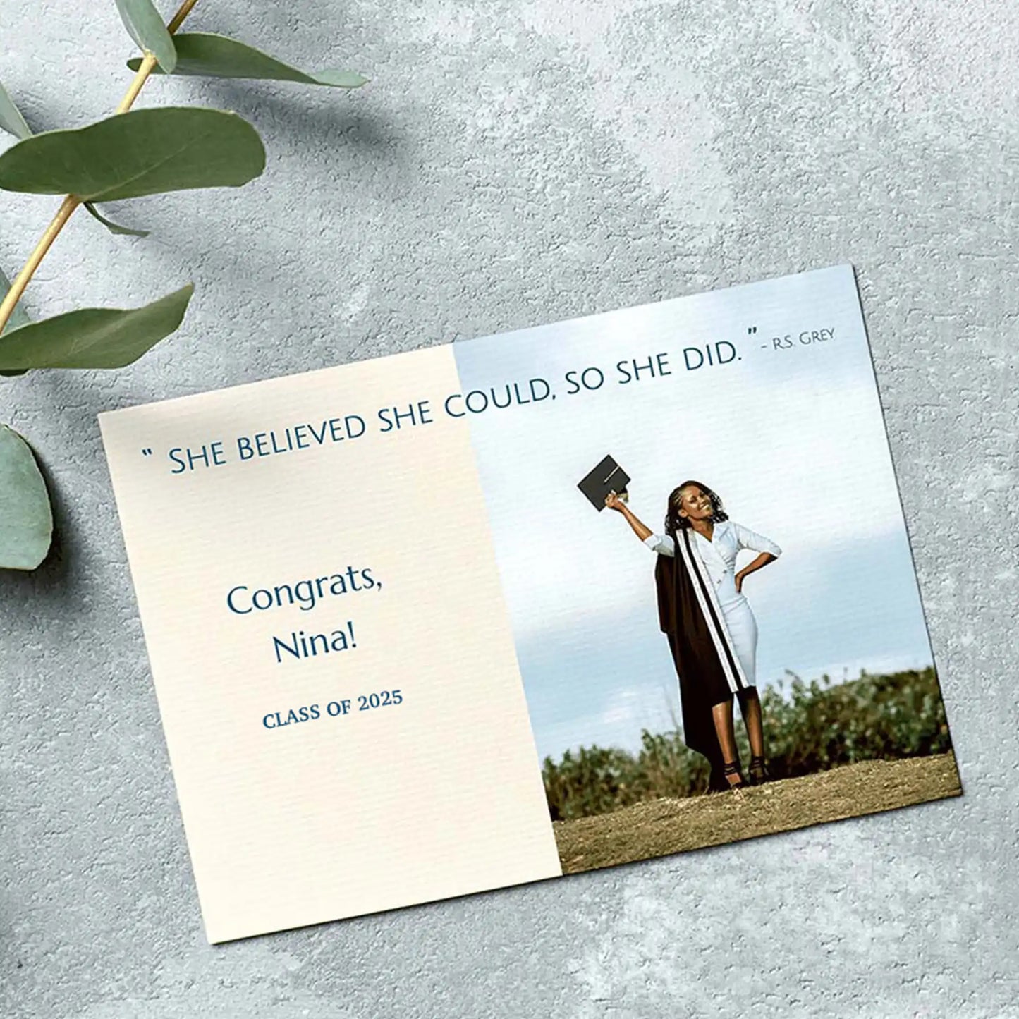 Flat graduation announcement card with inspirational quote and graduate holding cap outdoors on textured gray surface.