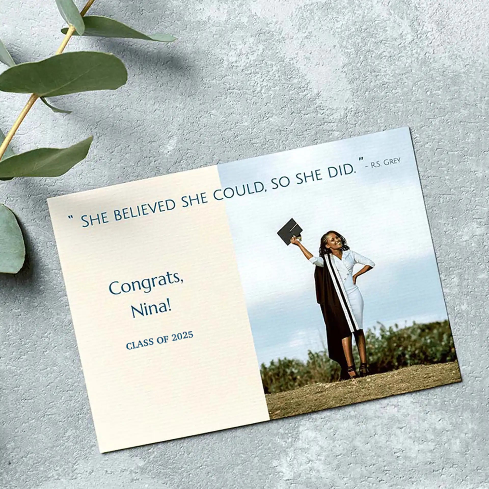 Flat graduation announcement card with inspirational quote and graduate holding cap outdoors on textured gray surface.