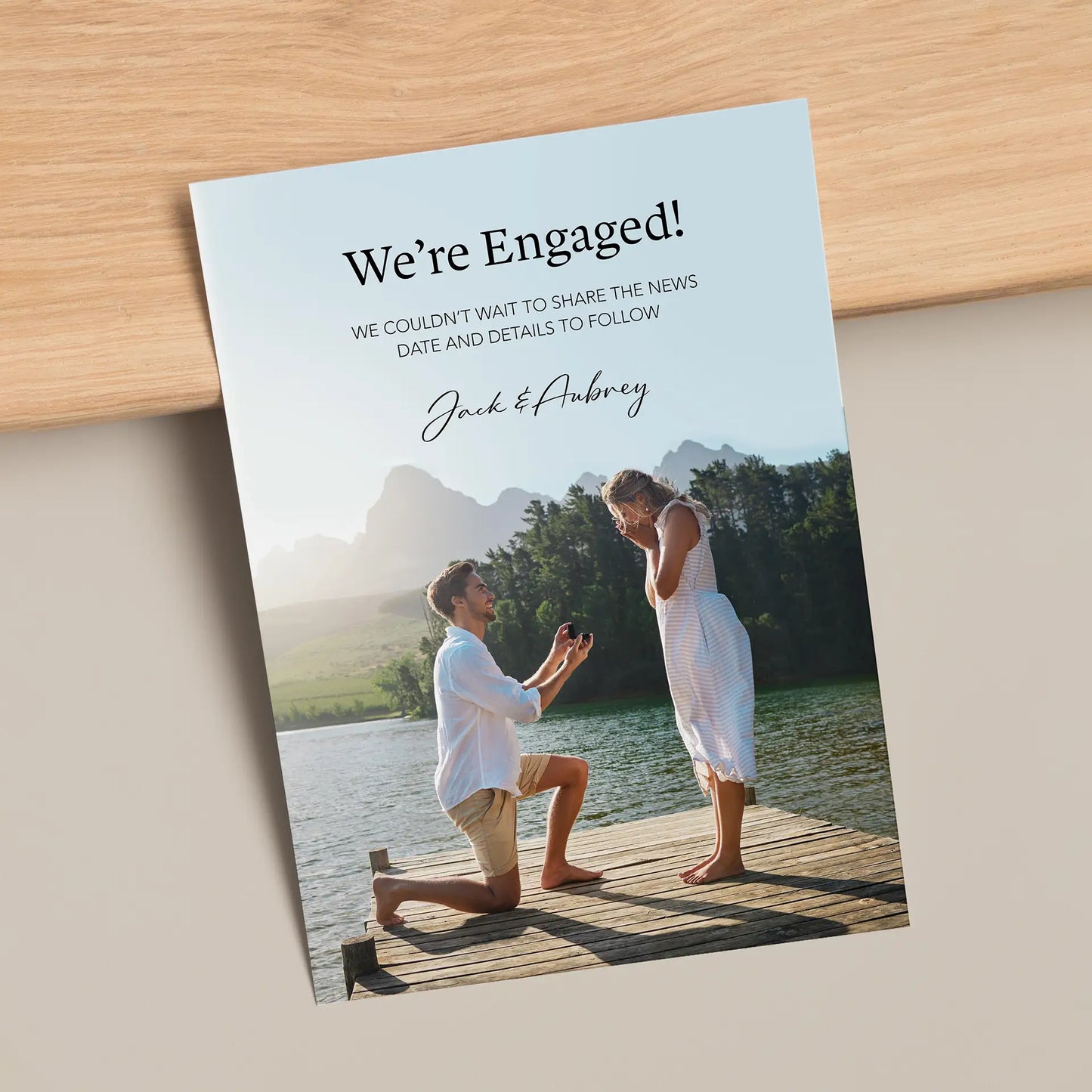 Flat engagement announcement card featuring lakeside proposal photo with modern typography on light wood surface.