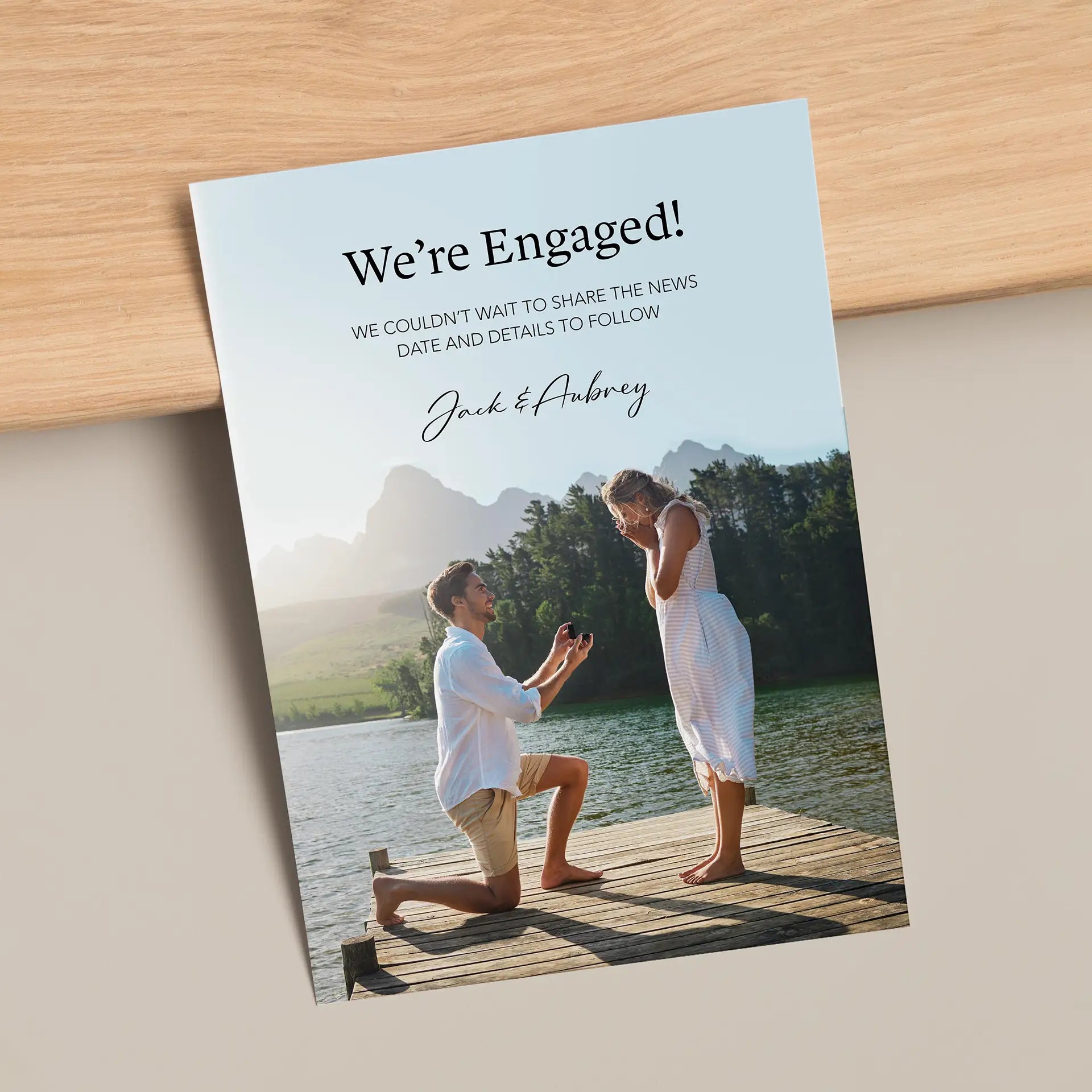 Flat engagement announcement card featuring lakeside proposal photo with modern typography on light wood surface.