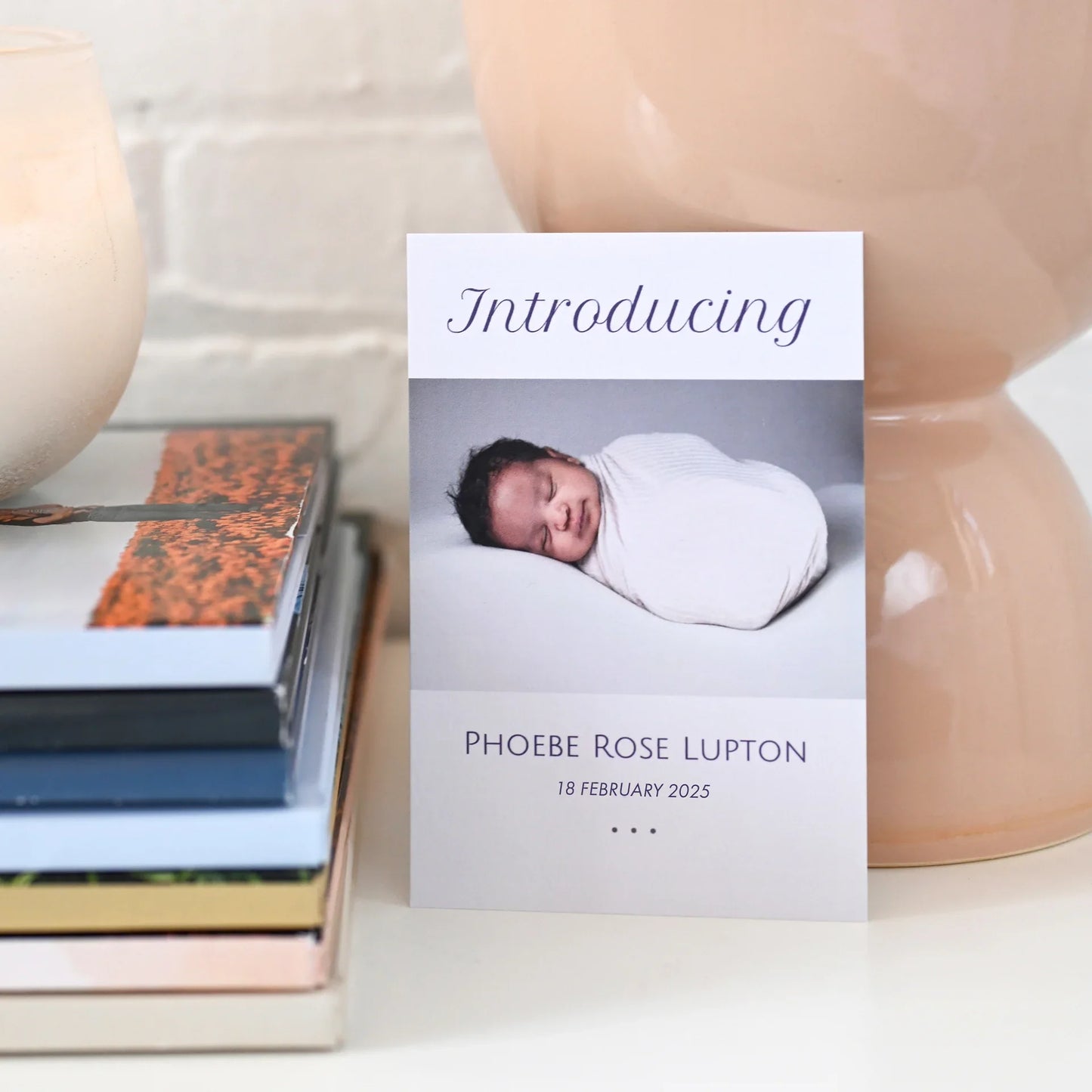 Flat birth announcement card with swaddled newborn portrait and name, displayed beside stacked books and ceramic vase.