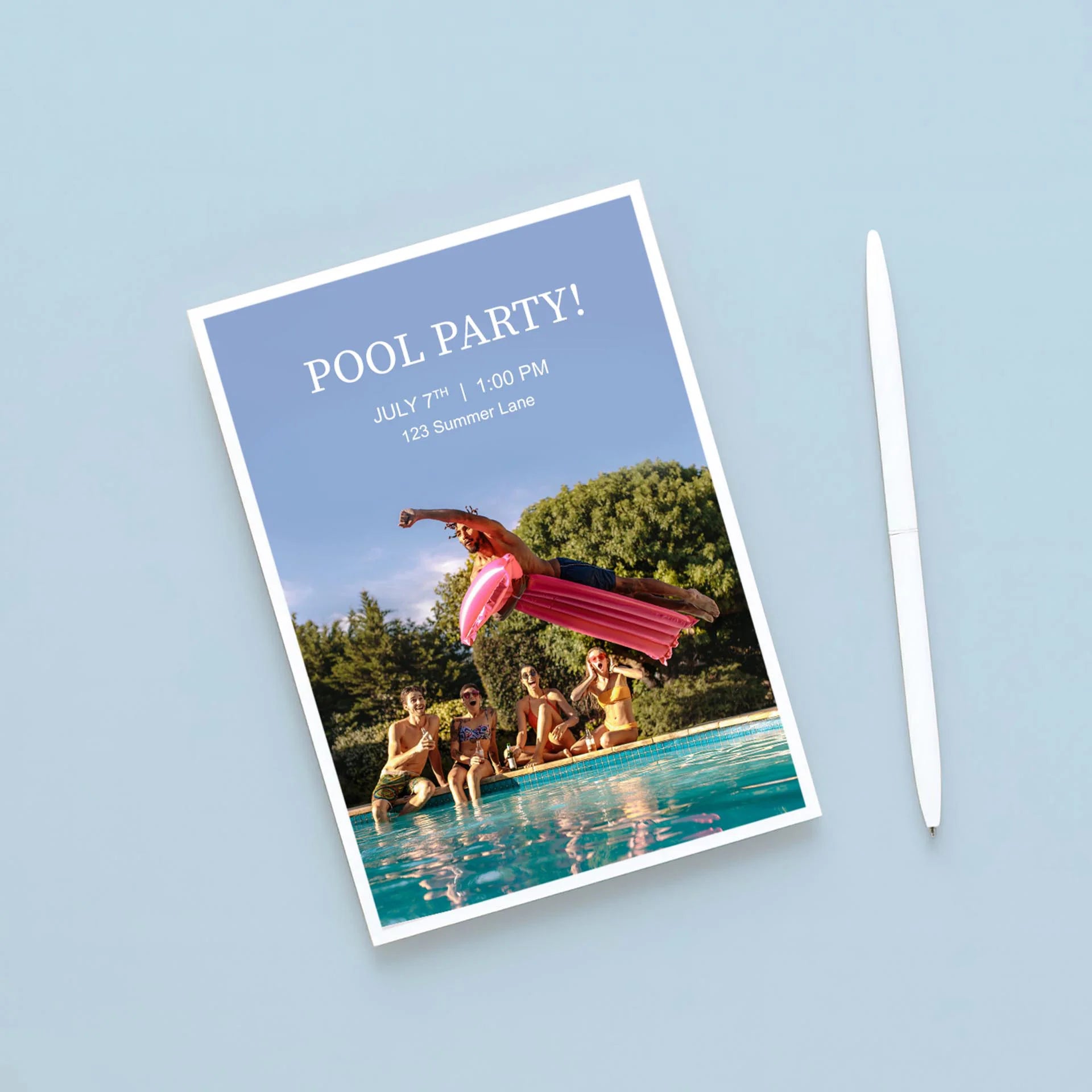 Flat invitation card with pool party text and photo of people by a swimming pool, placed on blue surface next to a white pen.