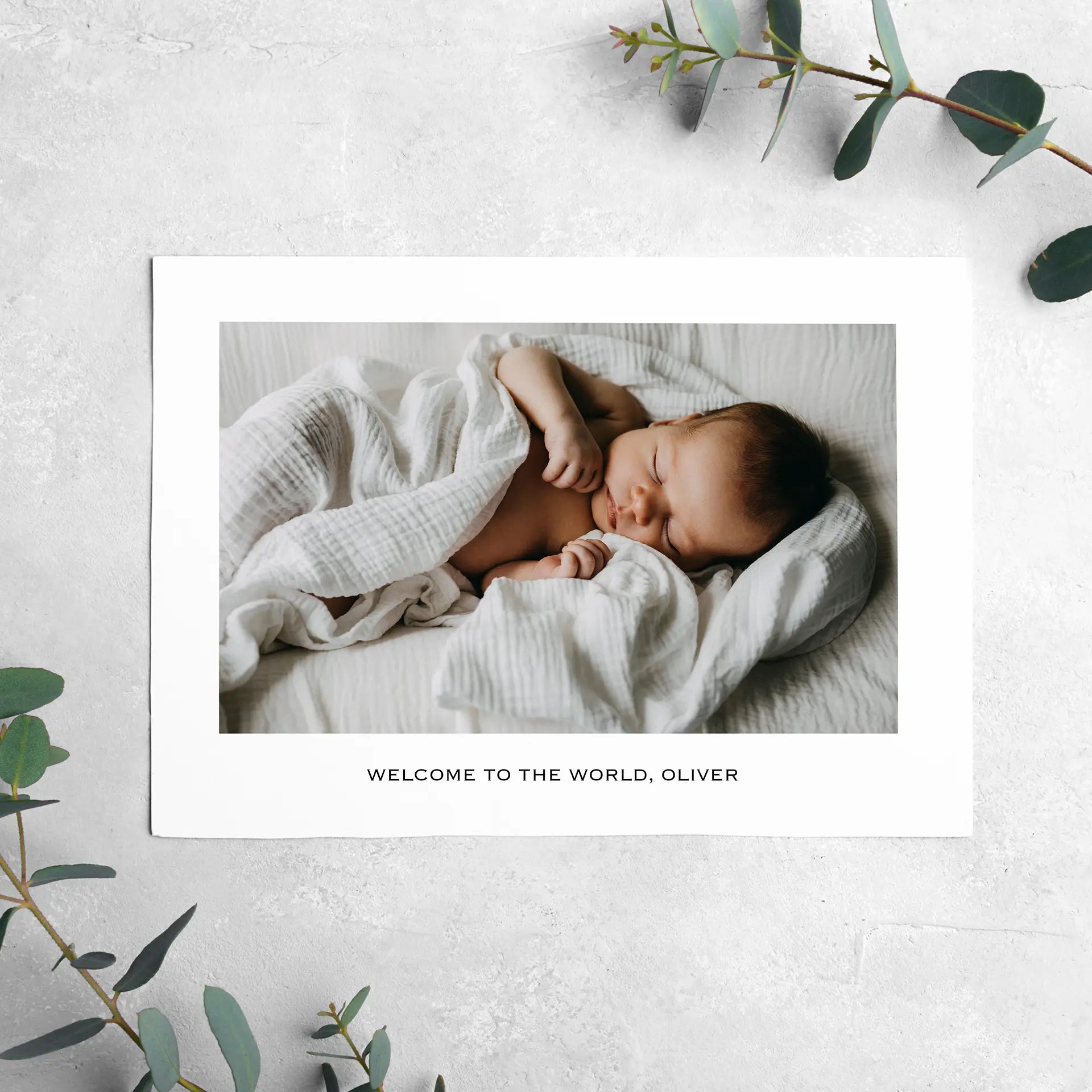 Flat landscape birth announcement card with sleeping newborn wrapped in white blanket on light textured background.