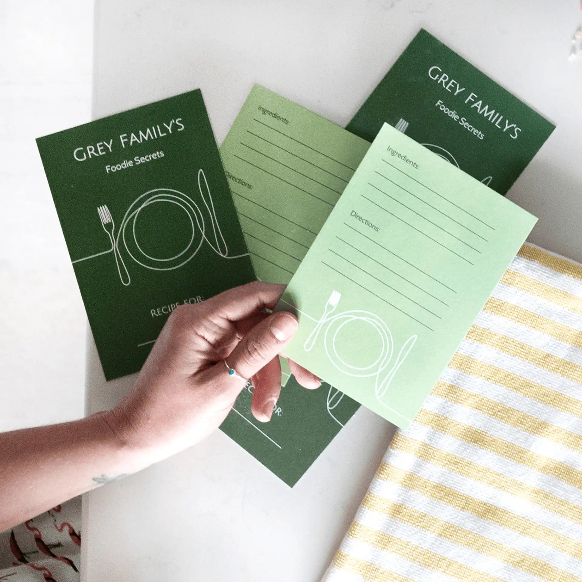 Set of green flat recipe cards with lined sections for ingredients and directions, held in hand beside striped cloth on a light surface.