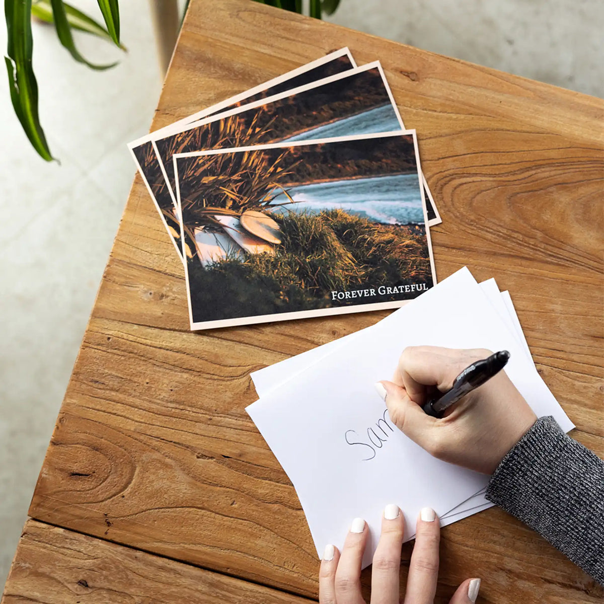 Stack of 'Forever Grateful' thank you flat cards with a beach sunset photo, beside a hand addressing an envelope.