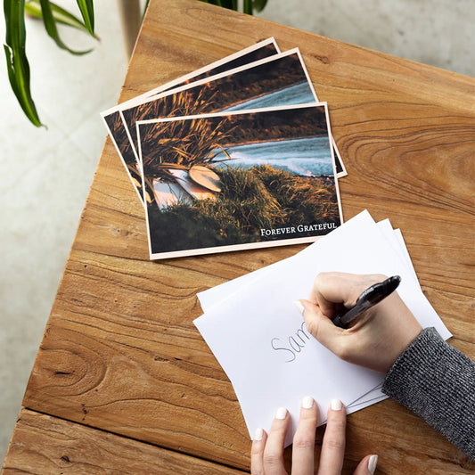 Stack of 'Forever Grateful' thank you flat cards with a beach sunset photo, beside a hand addressing an envelope.