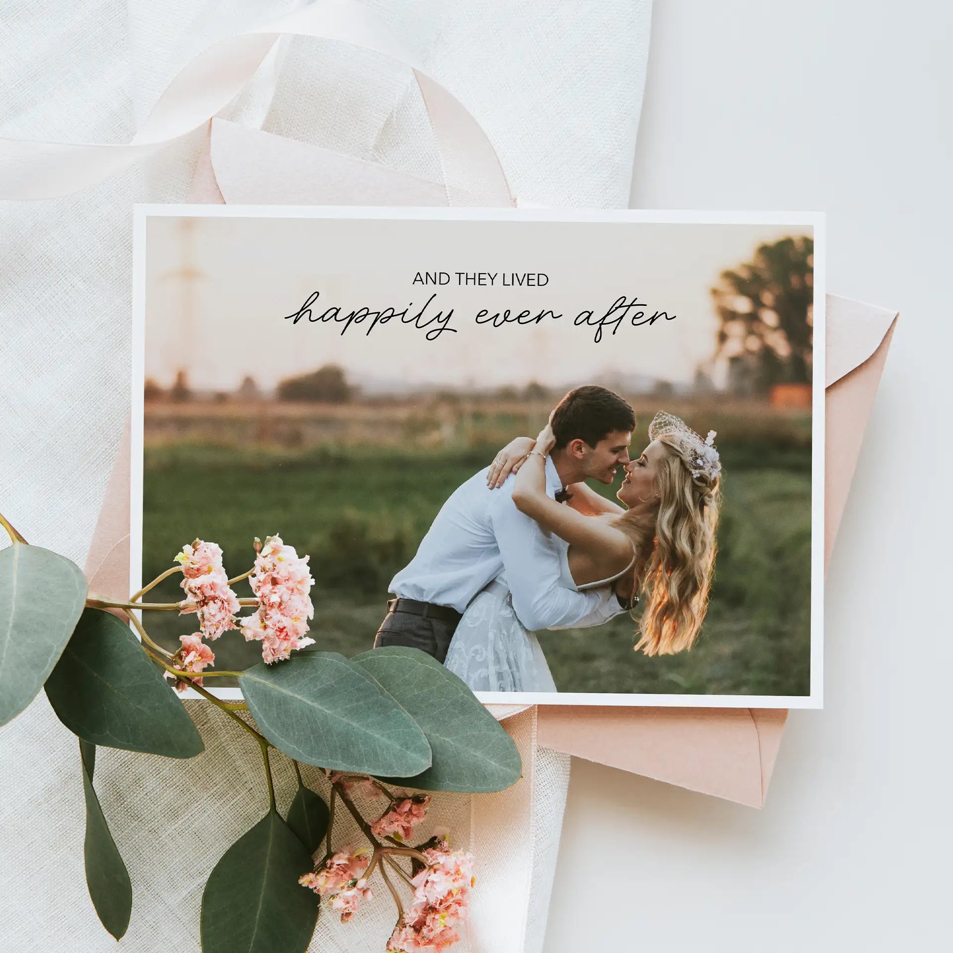 Folded wedding photo card featuring couple embracing at sunset in grassy field with script text reading “Happily Ever After.”