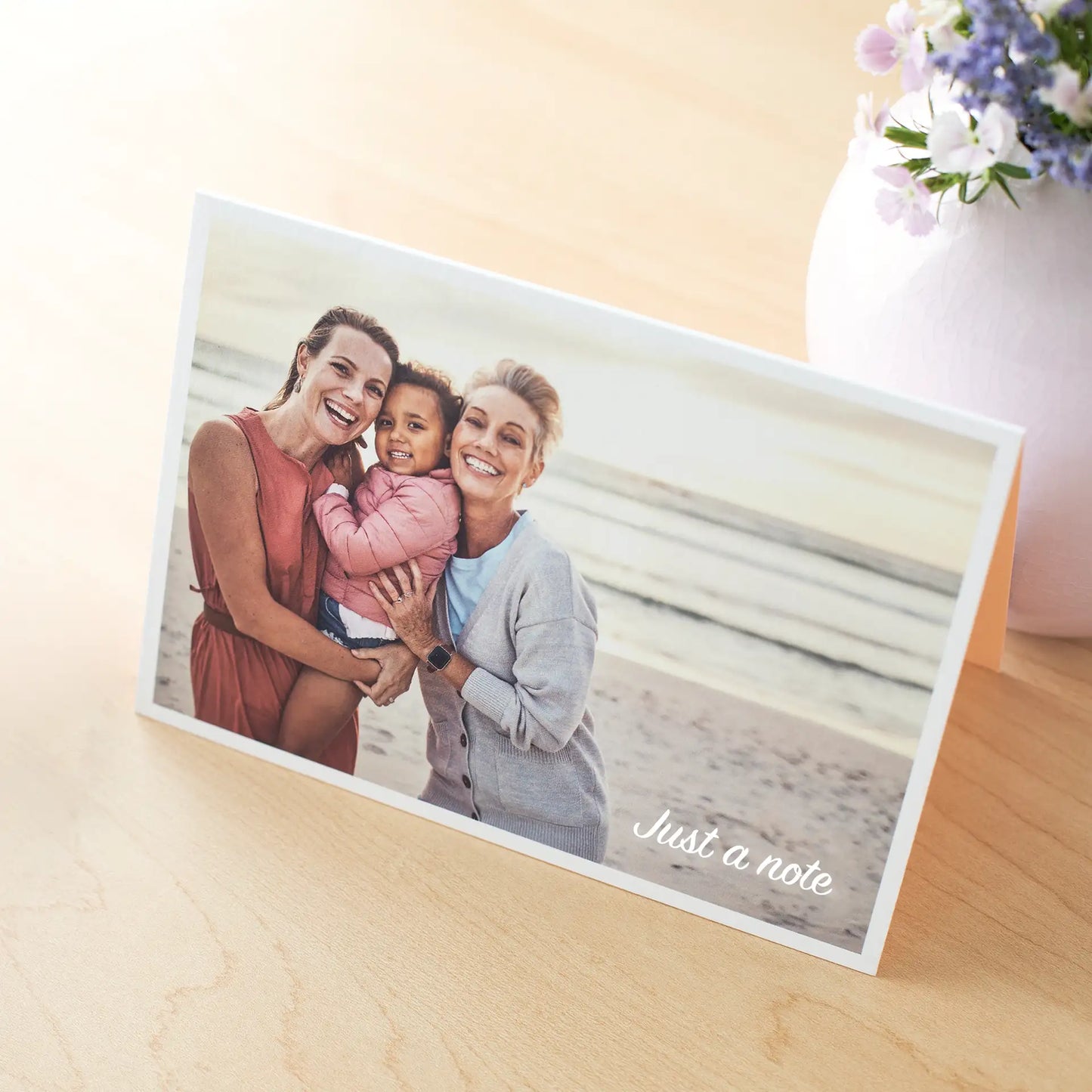 Folded card with family beach photo of two adults and a child smiling together, “Just a note” text visible, placed on wooden surface near vase of flowers.