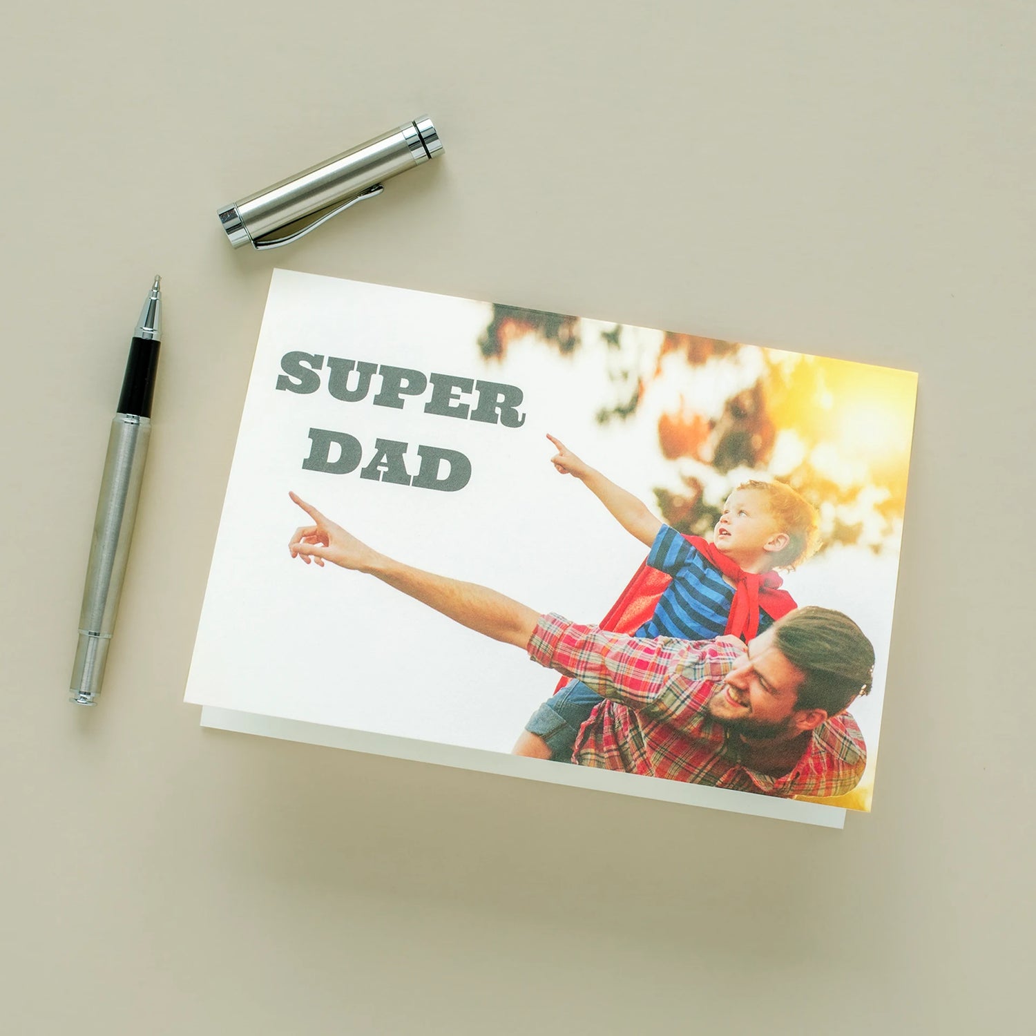Custom folded Father's Day card reading 'Super Dad' lying on a neutral surface beside two pens, featuring a photo of a father smiling as his young son in a superhero cape sits on his shoulders pointing into the sky