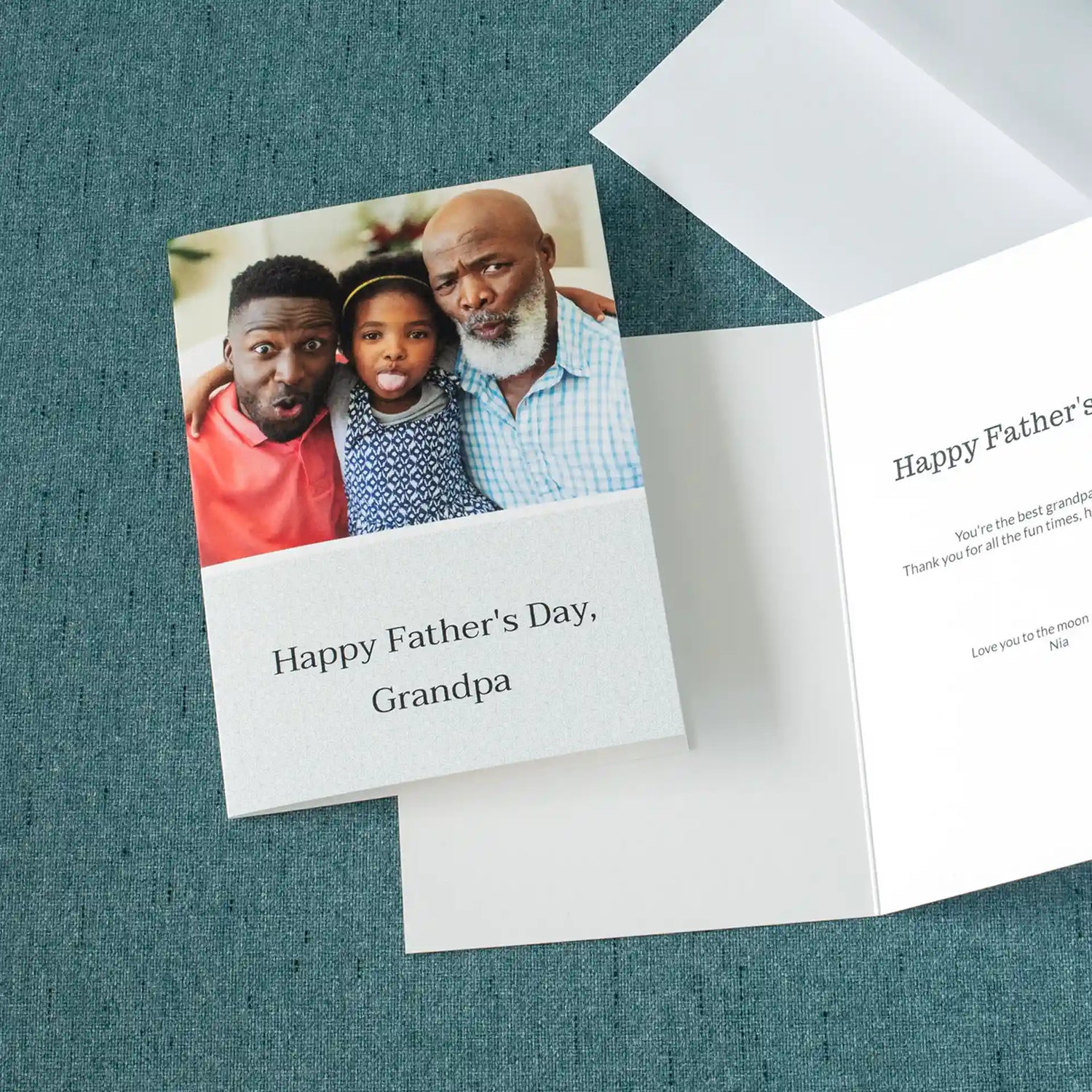 Custom folded Father's Day card reading 'Happy Father's Day, Grandpa' shown front and open beside its envelope on a teal surface, featuring a playful photo of a grandfather, father, and young girl making silly faces together