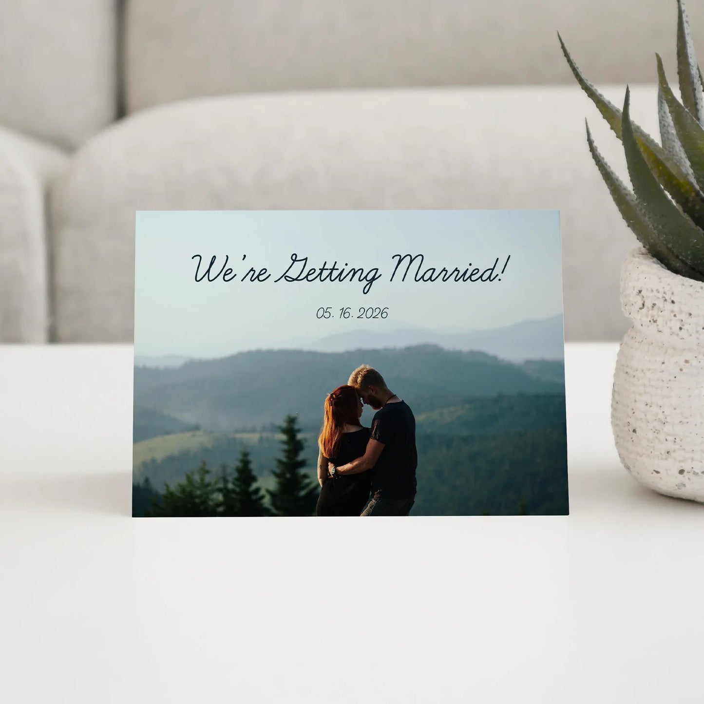 Folded wedding announcement card featuring couple embracing against mountain landscape with printed date and script headline above photo.