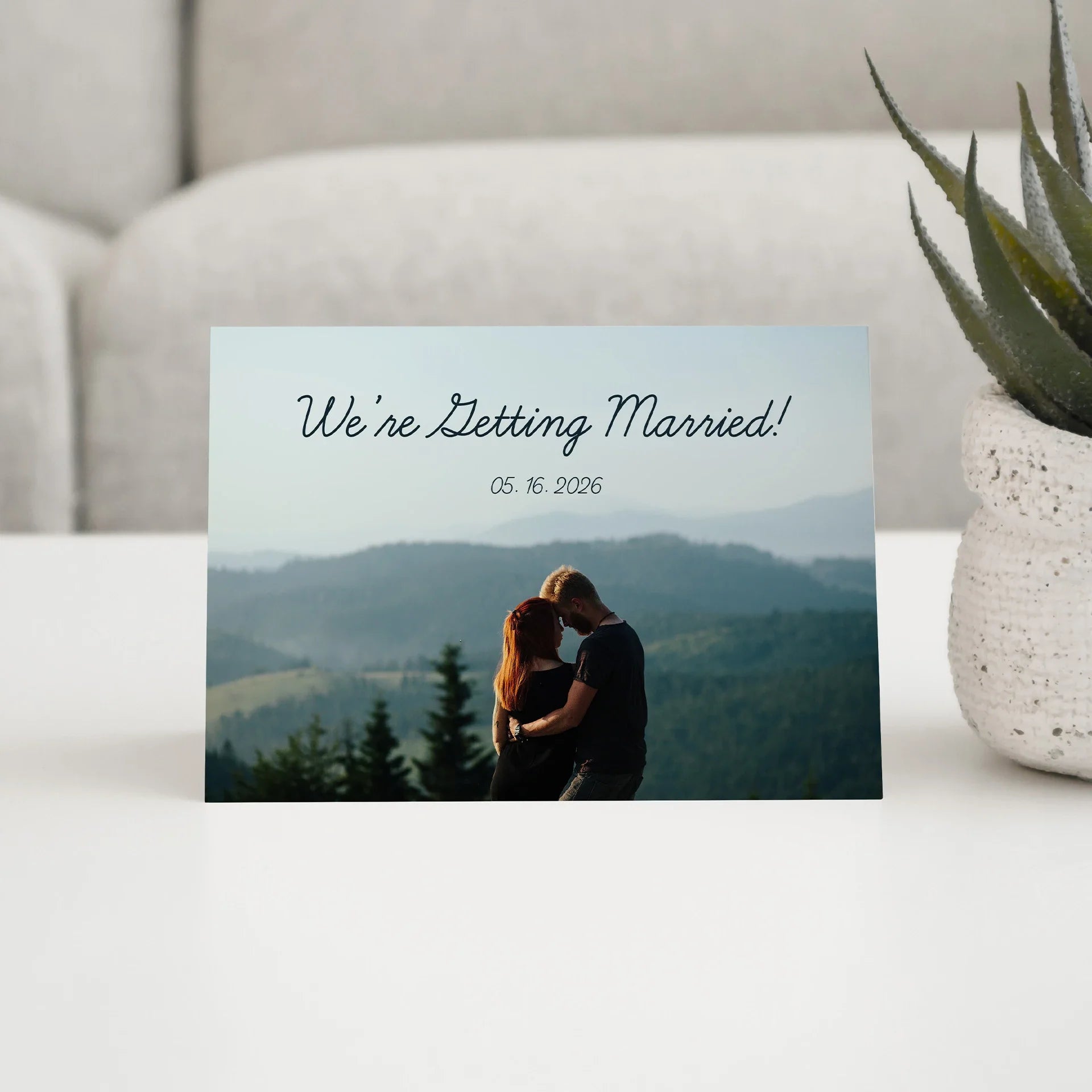 Folded wedding announcement card featuring couple embracing against mountain landscape with printed date and script headline above photo.
