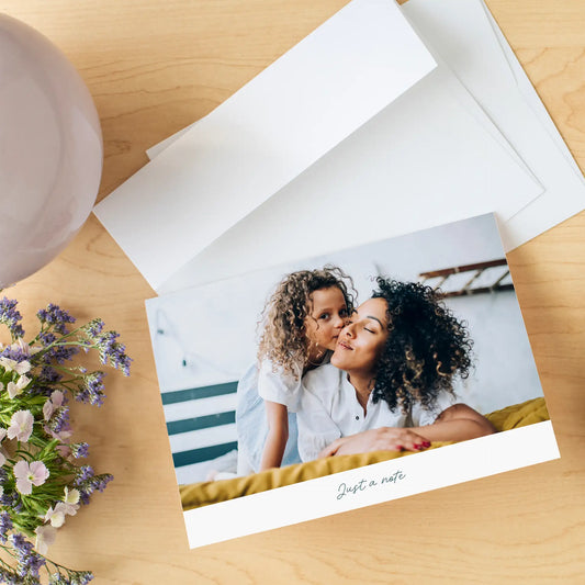 Folded photo card with image of a child hugging a person, “Just a note” text, styled with envelope and flowers on wood surface.