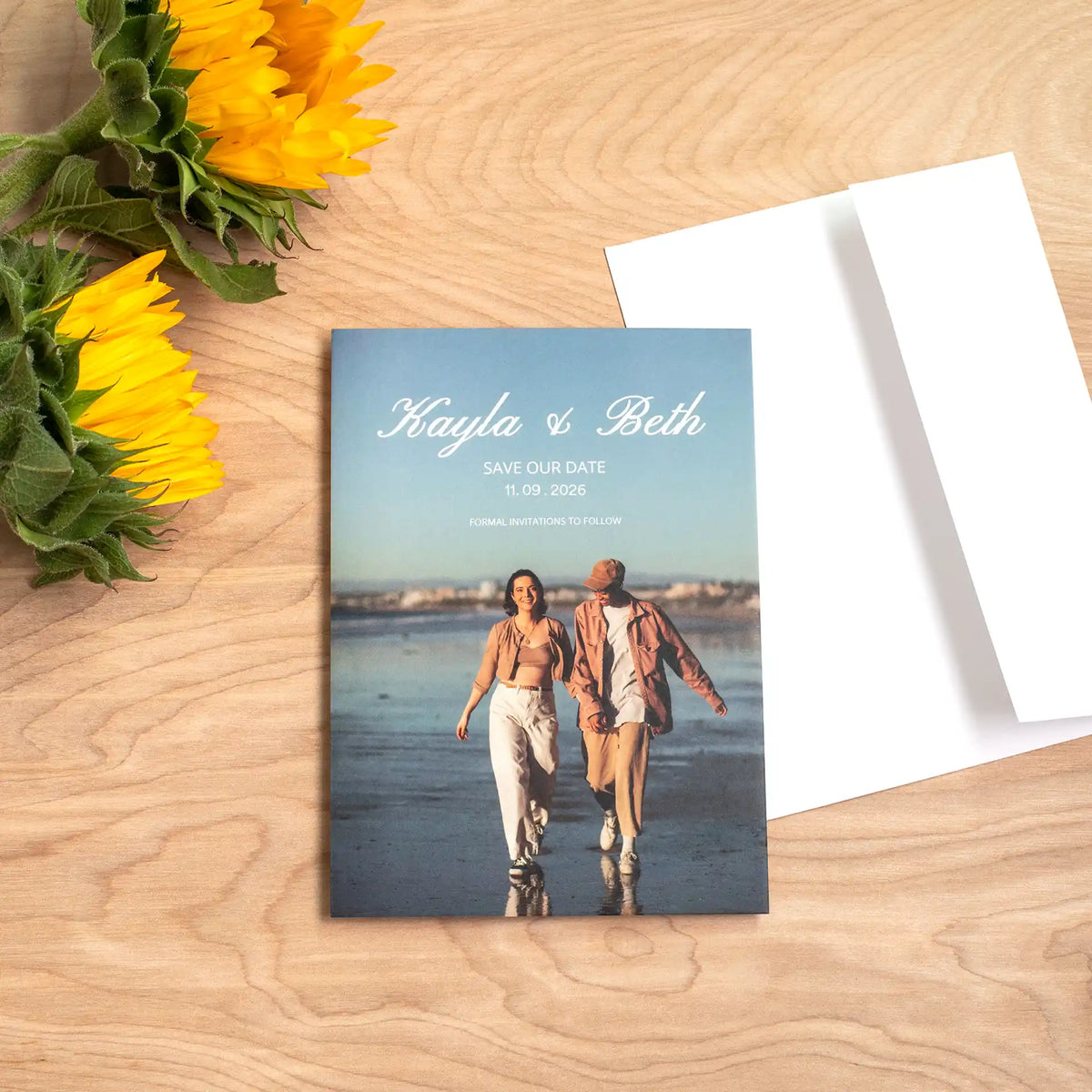 Folded save-the-date card featuring two women walking along a beach, reading 'Kayla & Beth, Save Our Date,' beside sunflowers