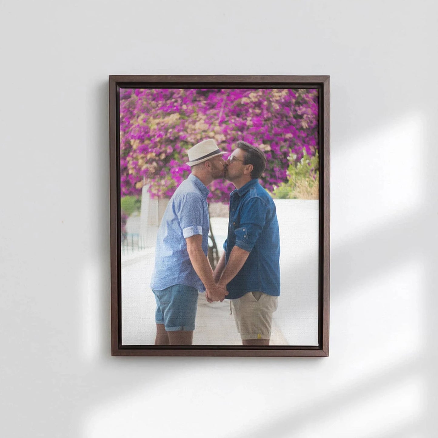 Dark wood framed canvas print of two men holding hands and kissing beneath vibrant pink flowering trees, mounted on a white wall.