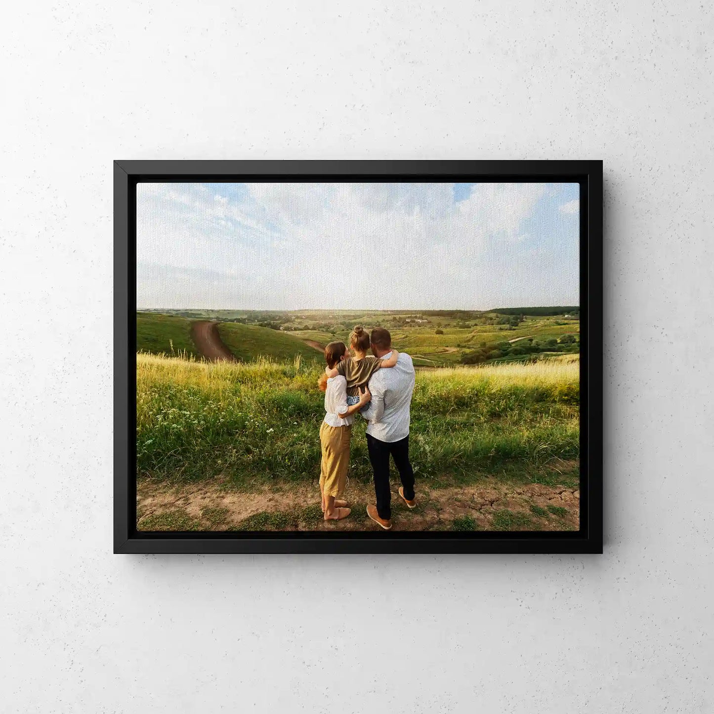 Framed canvas print with black frame showing family of three from behind overlooking rolling green hills, landscape orientation, displayed on light textured wall.
