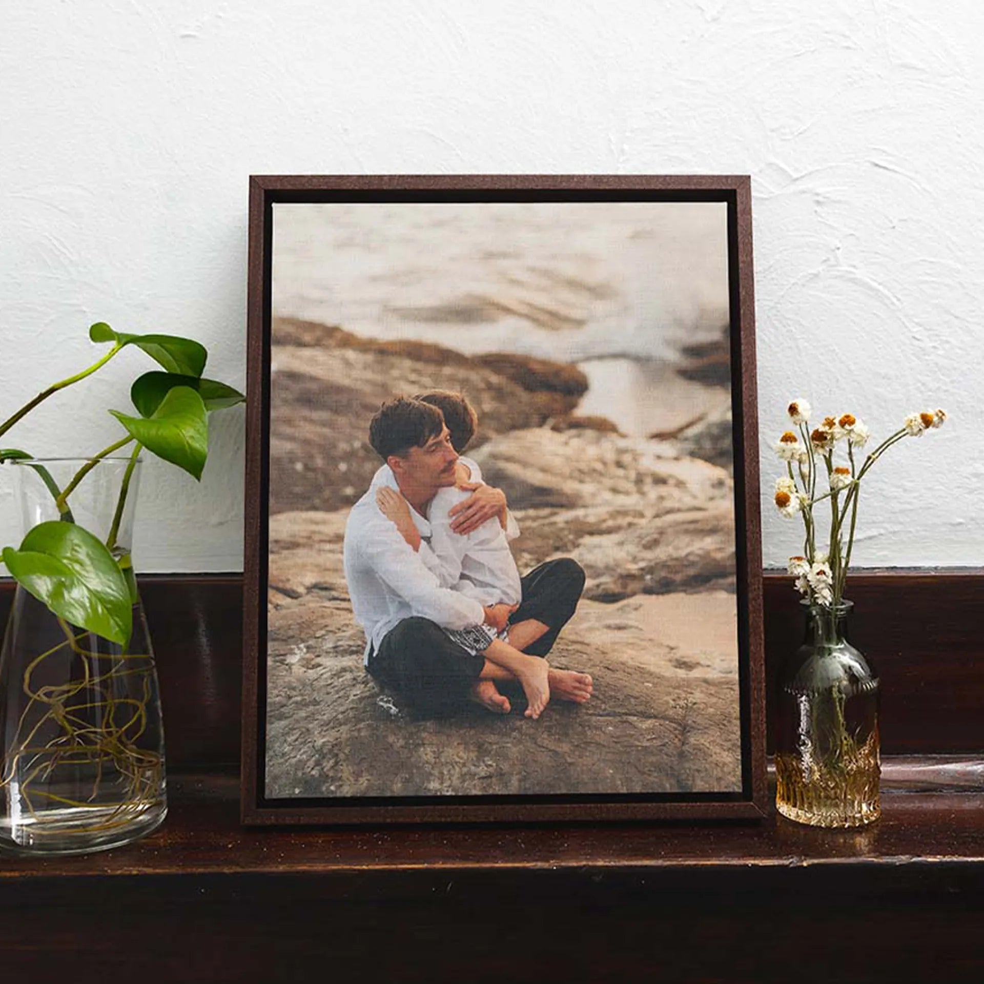 Dark wood framed canvas featuring a father sitting on rocks hugging his child near the shoreline, styled on a wooden ledge with plants.