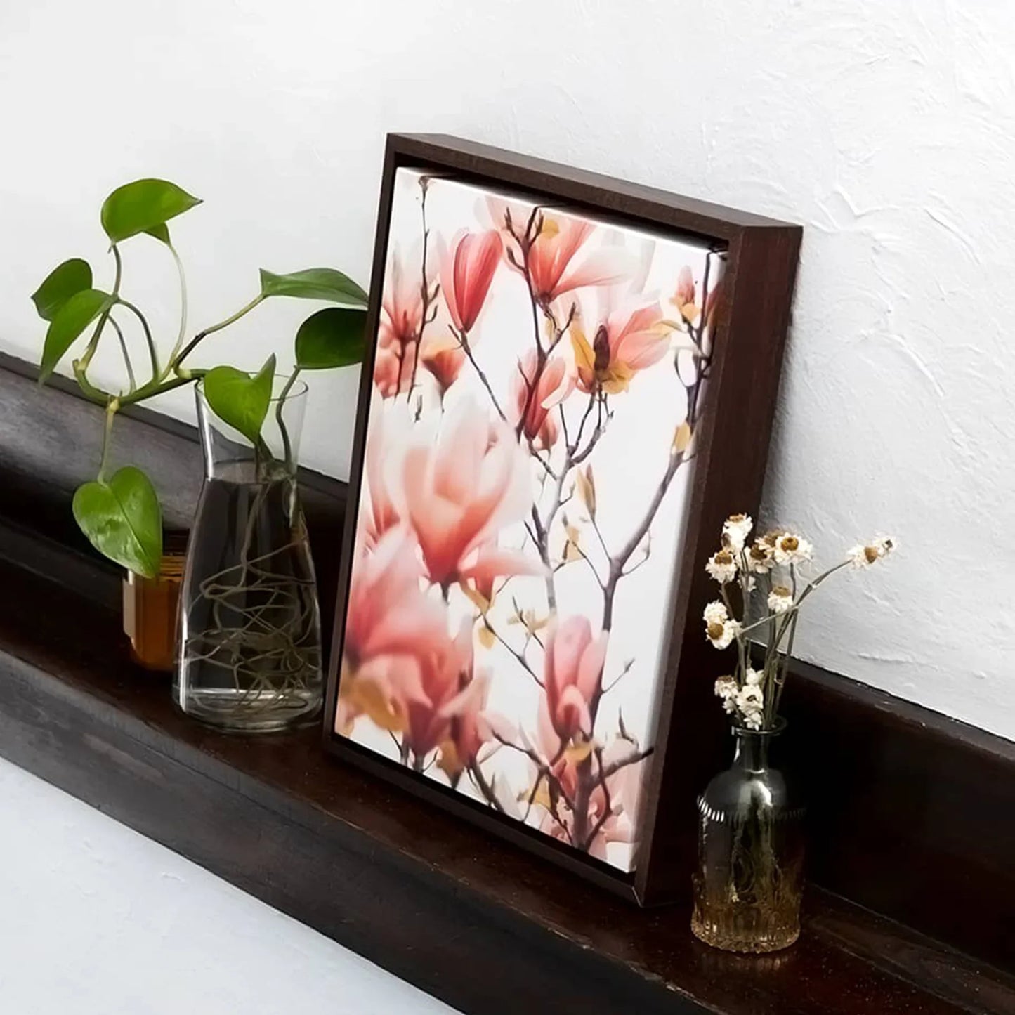 Dark wood framed canvas featuring pink magnolia blossoms leaning on a wooden shelf beside glass vases with greenery.