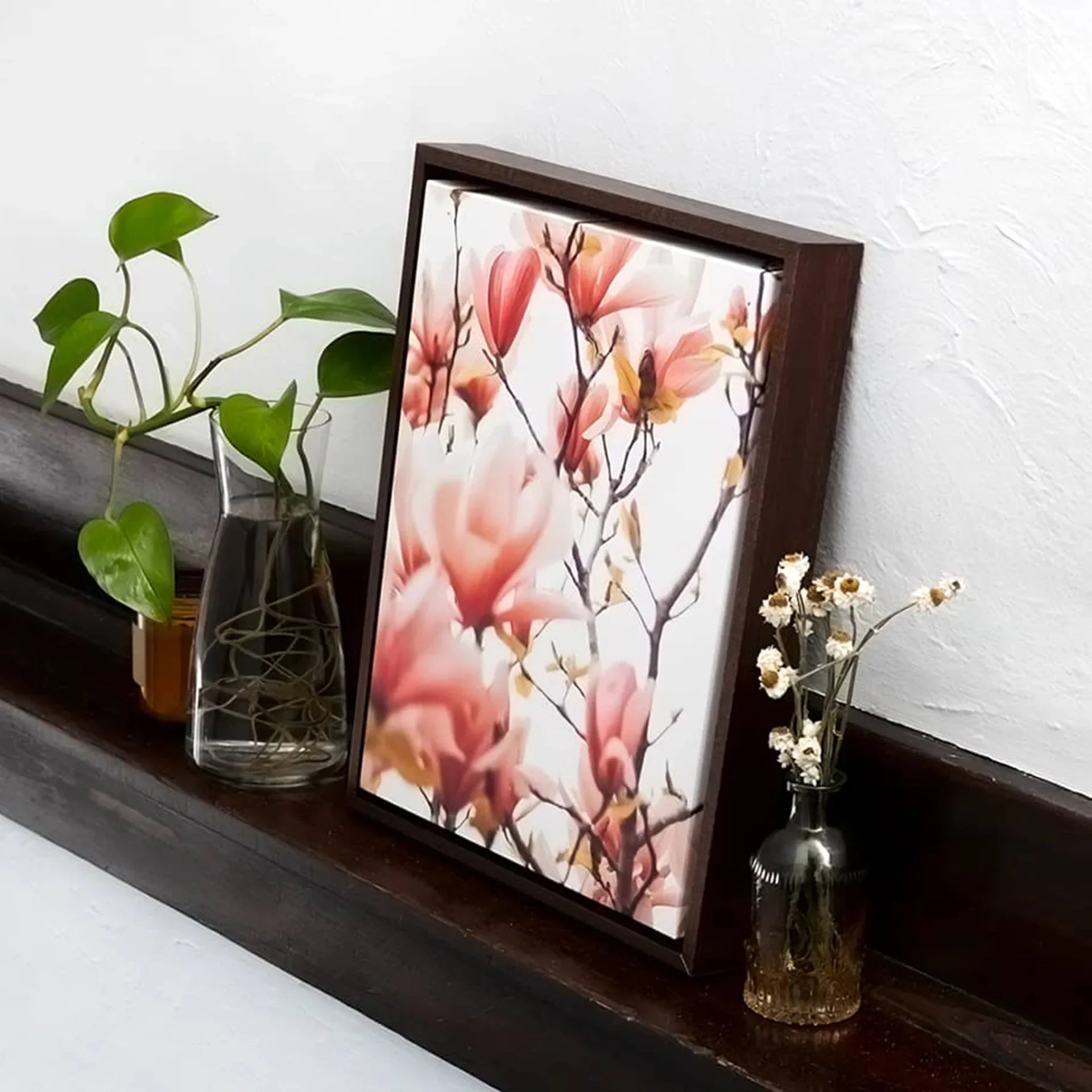 Dark wood framed canvas featuring pink magnolia blossoms leaning on a wooden shelf beside glass vases with greenery.