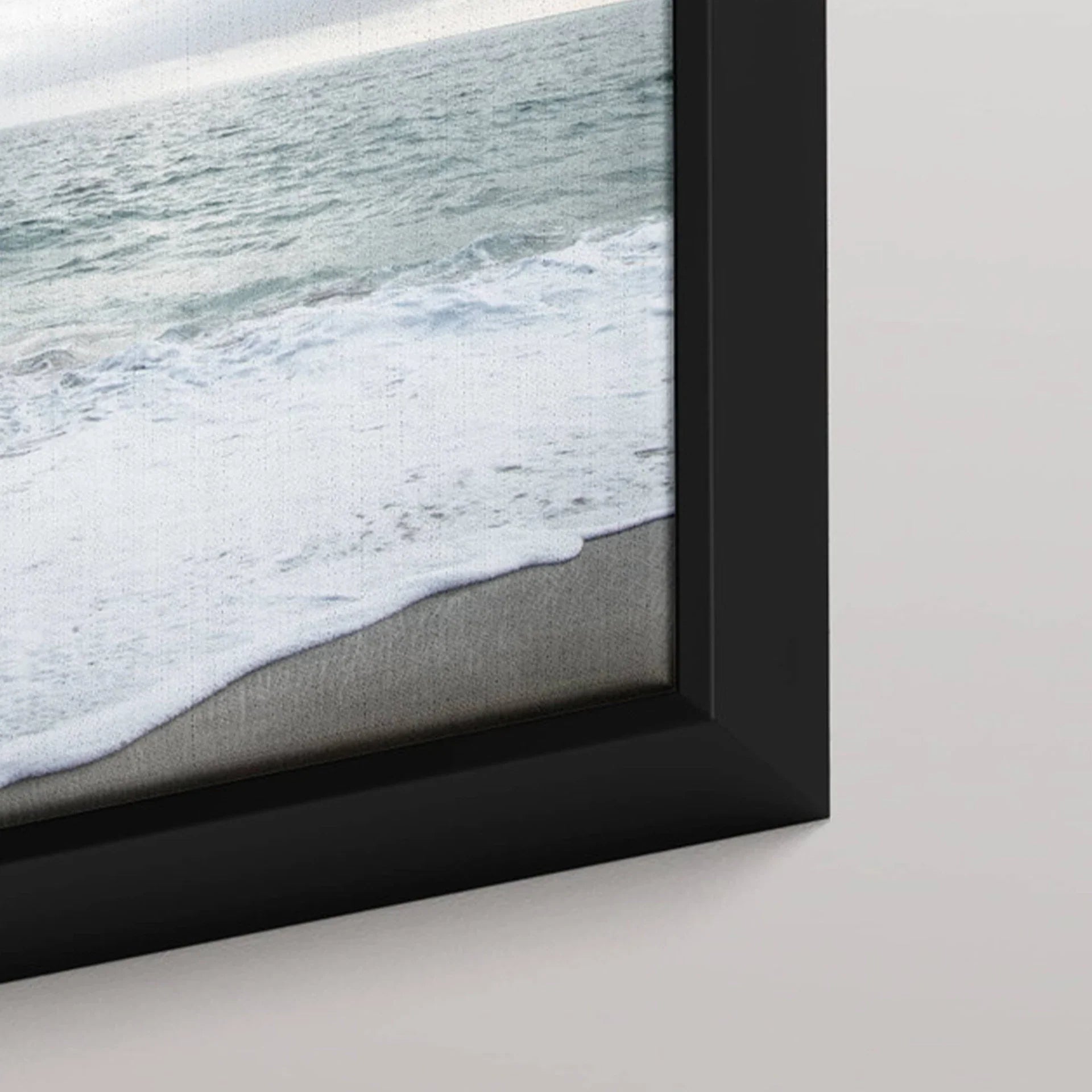 Black framed canvas print featuring ocean waves rolling onto sandy shore, shown in close-up detail against a light wall.