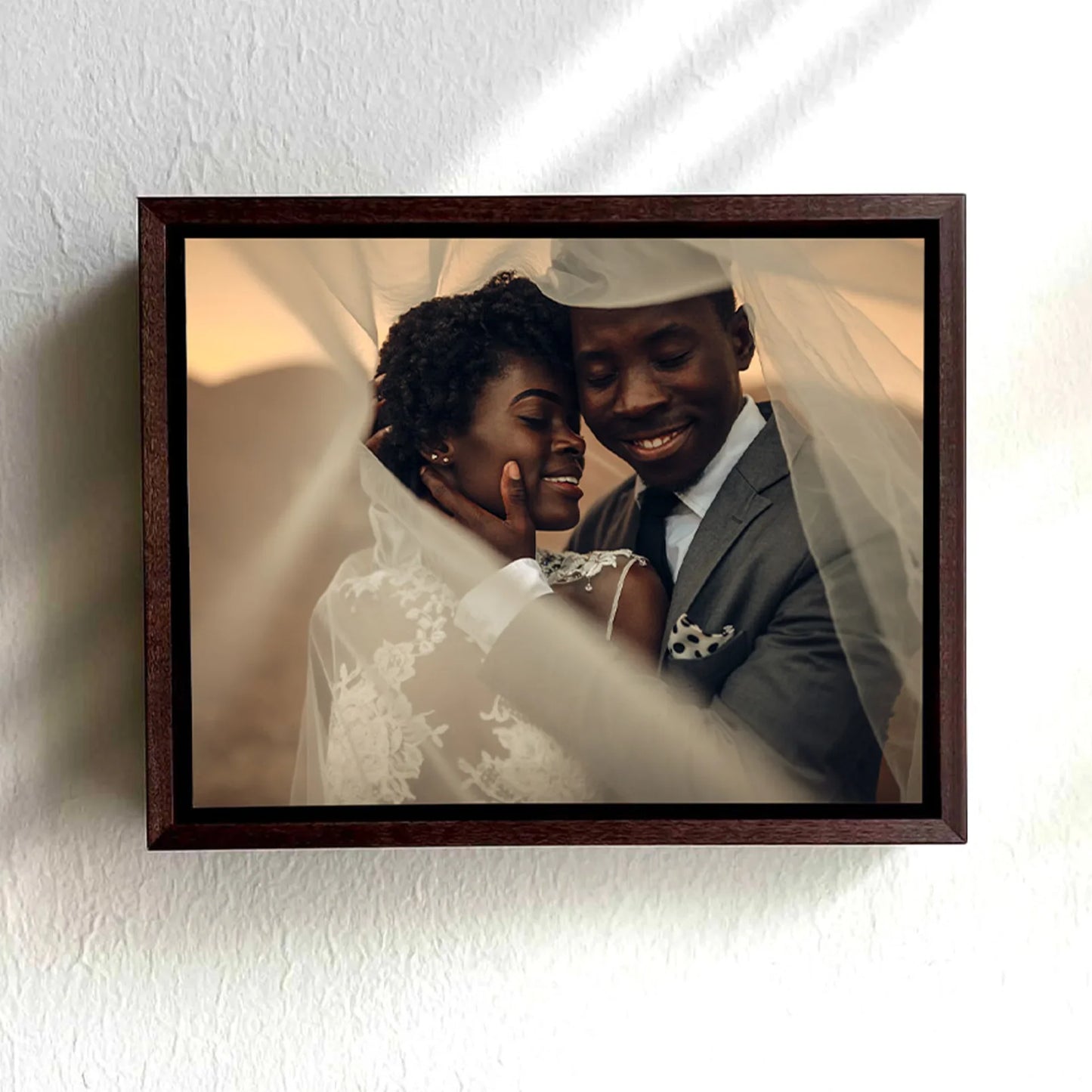 Dark wood framed canvas print of a bride and groom embracing beneath a flowing veil, mounted on a white wall.