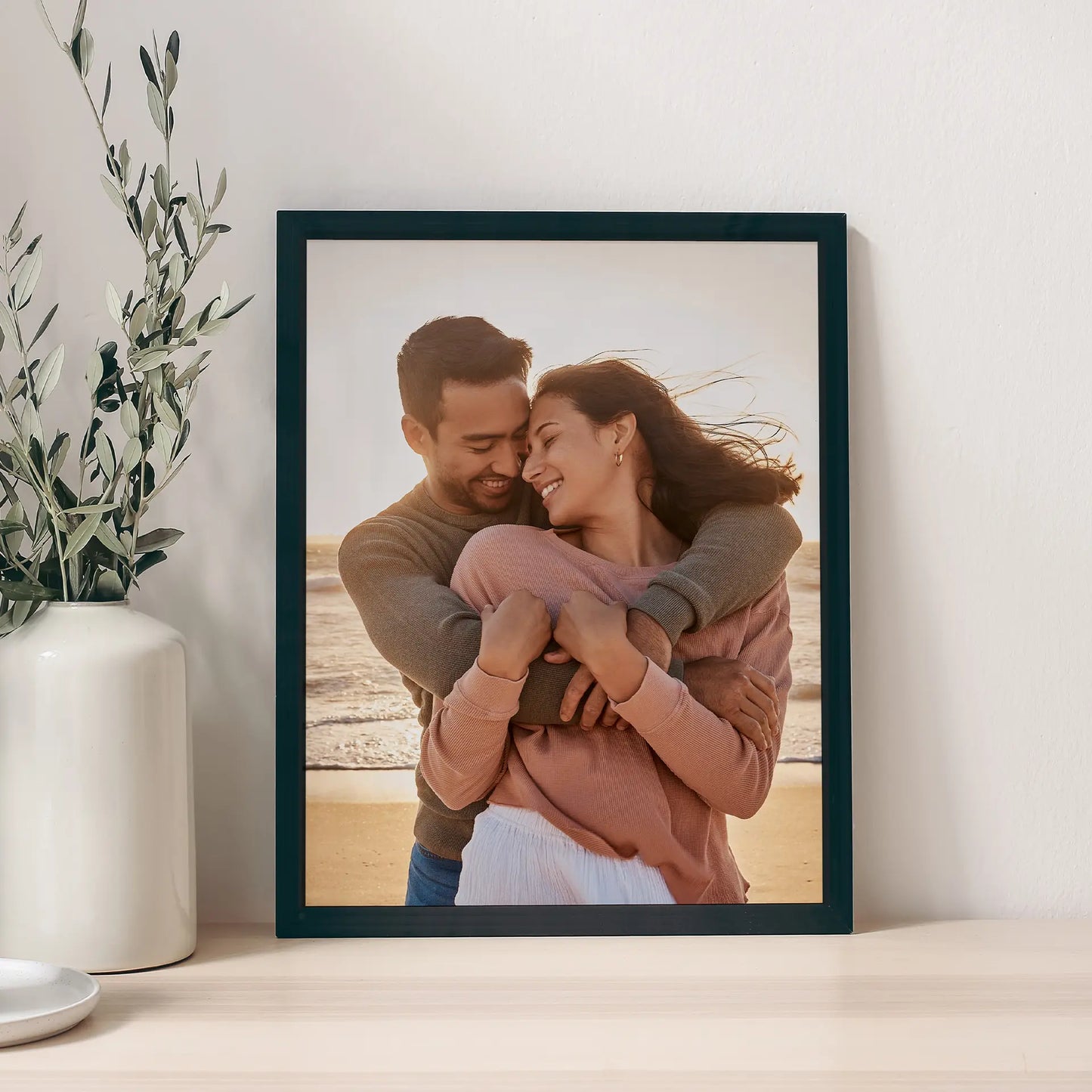 Black framed vertical photo print of a couple embracing and smiling at sunset on a beach, displayed on a tabletop beside a white vase with greenery.