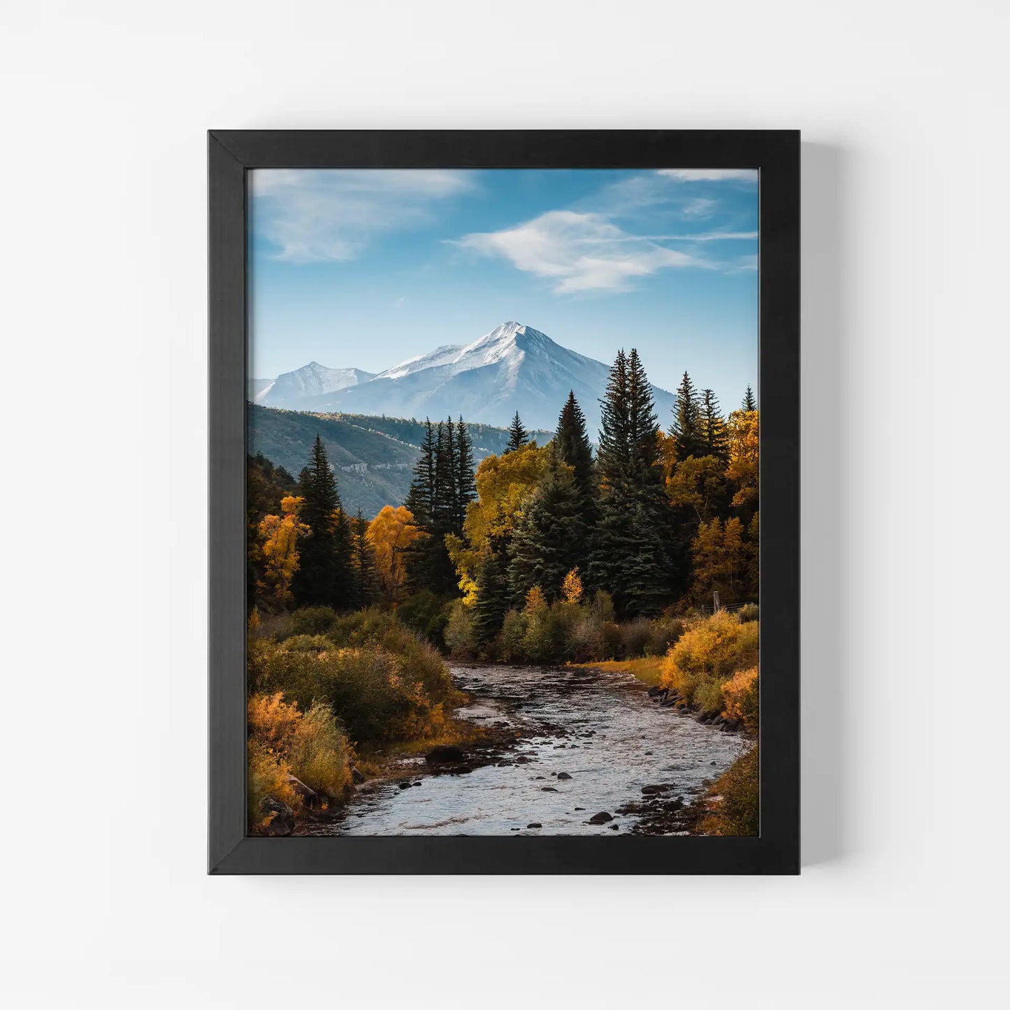 Framed print with black frame showing mountain landscape with river, evergreen trees, and autumn foliage, vertical orientation, displayed on light neutral wall.