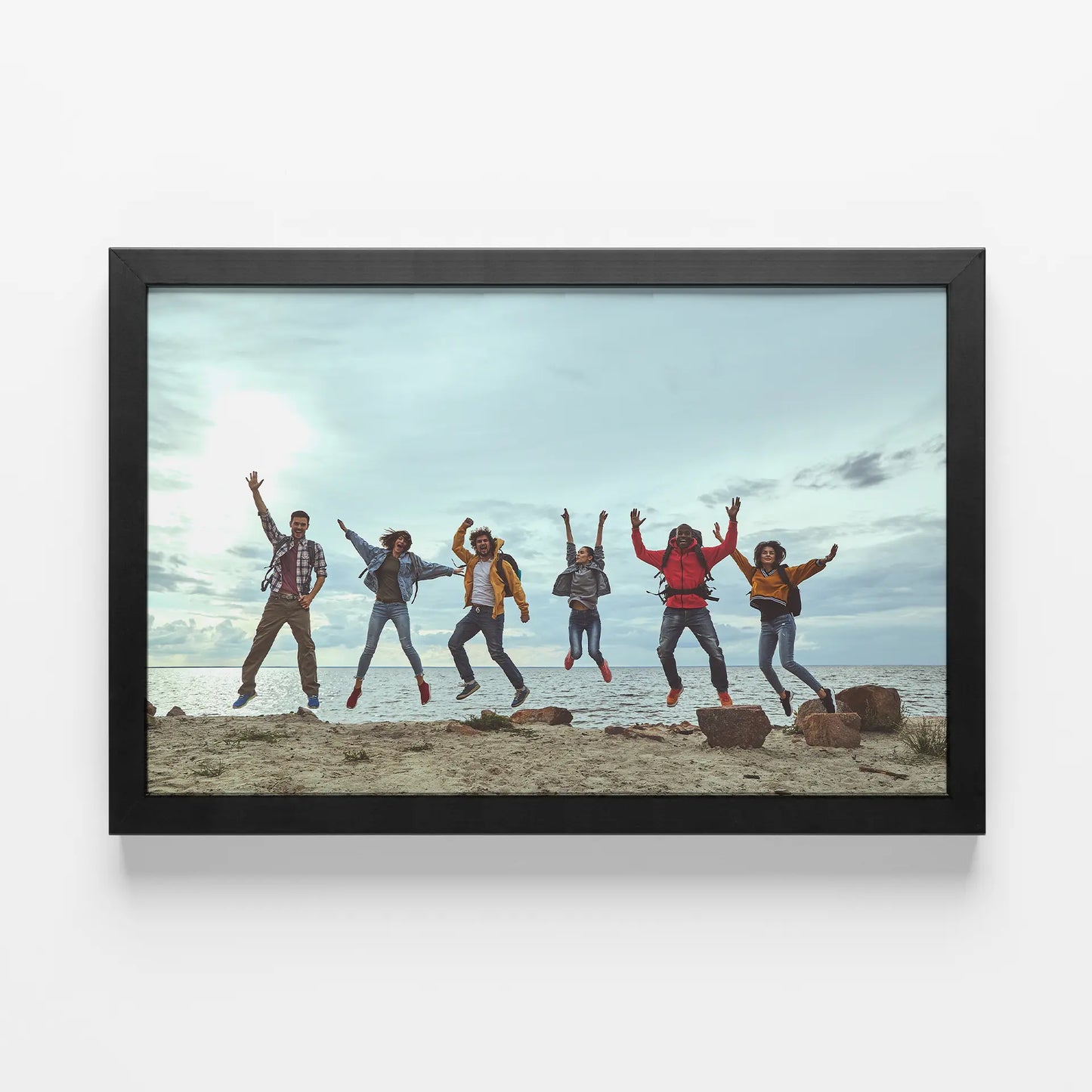 Framed print with black frame featuring group of people jumping on beach near ocean, wide landscape orientation, cloudy sky background, displayed on wall.