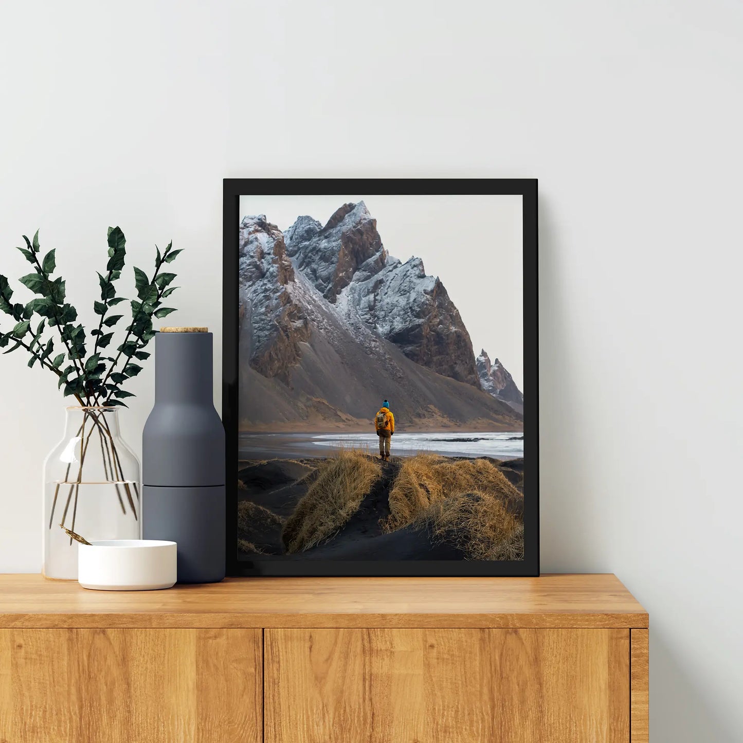 Black framed vertical photo print of a person in a yellow jacket standing on grassy dunes before snow-covered mountains and water, displayed on a wooden cabinet with decorative vases.