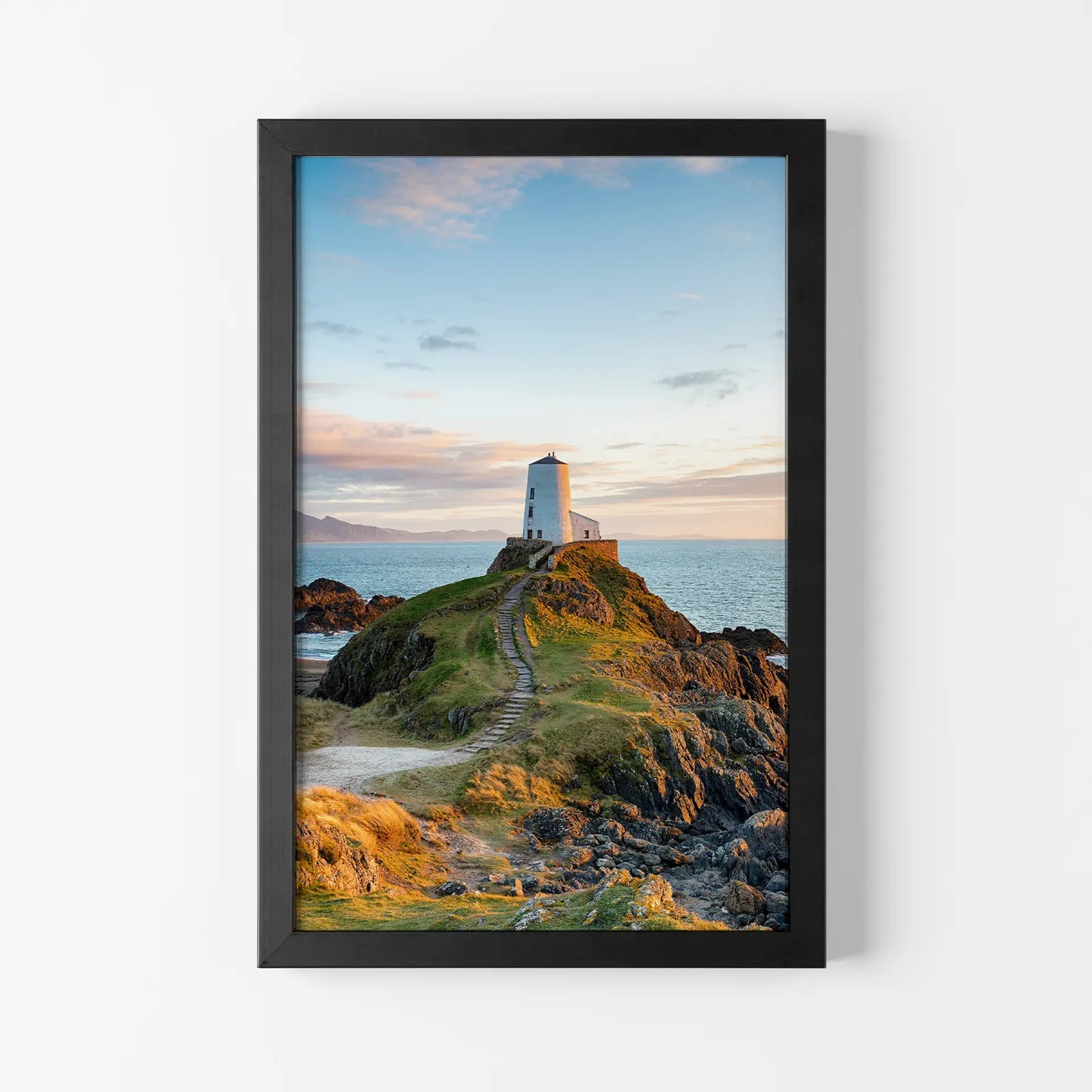 Framed print with black frame showing small white lighthouse on rocky coastal hill with path leading to it, vertical orientation, ocean and sky backdrop.