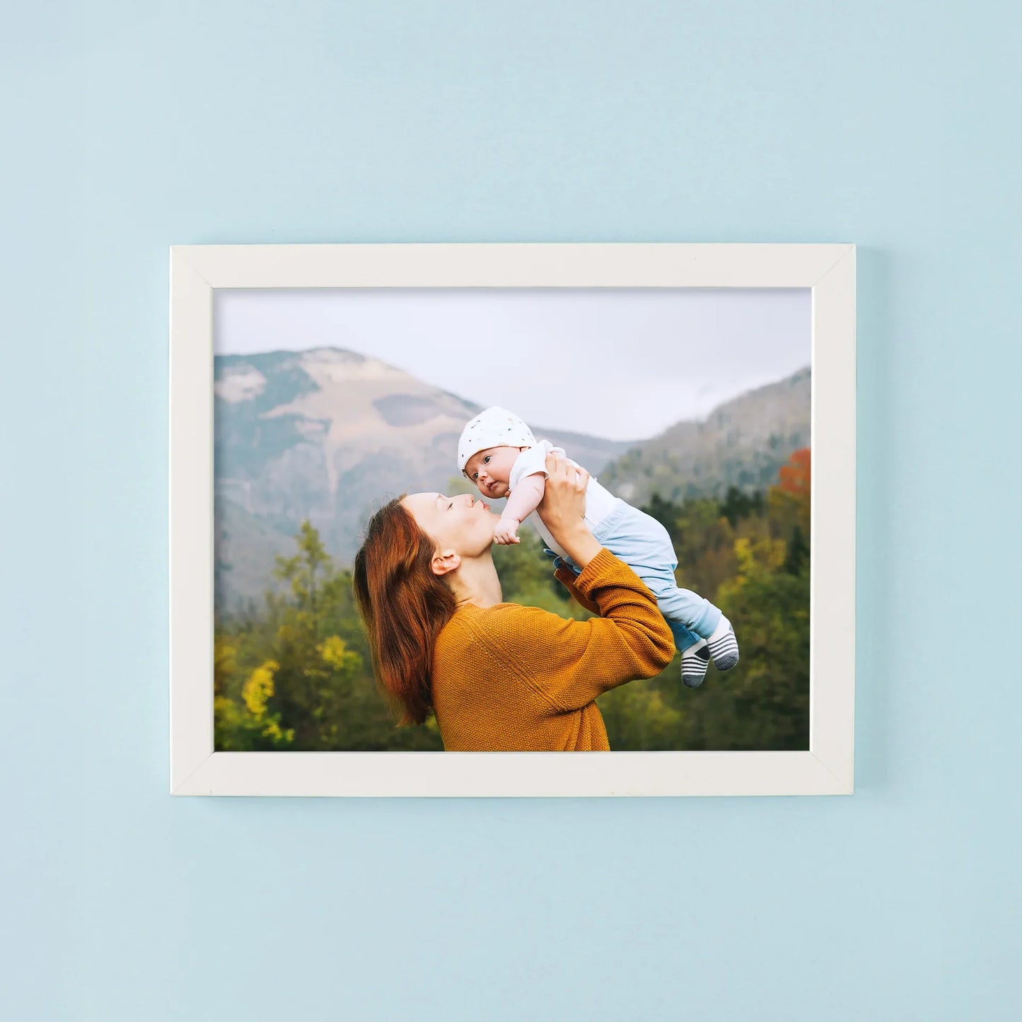 Framed print with white frame showing person lifting baby outdoors with mountains and trees in background, horizontal orientation, displayed on light blue wall.