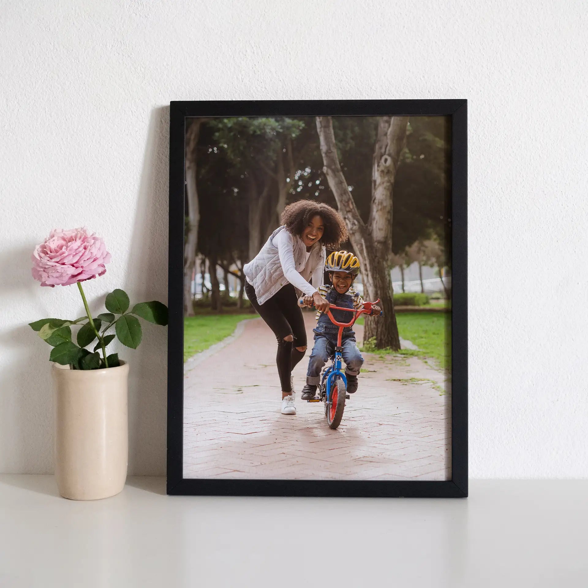 Black framed vertical photo print of a woman helping a child ride a bicycle on a paved park path, displayed on a shelf.