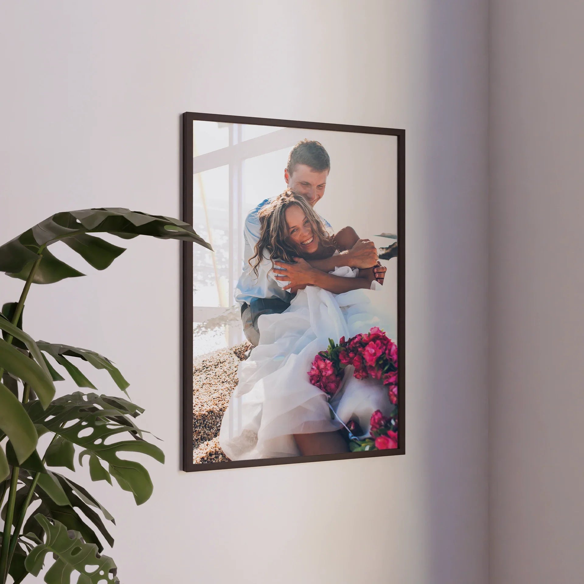 Dark wood framed vertical photo print of a bride and groom embracing near a window with pink flowers visible in the foreground.
