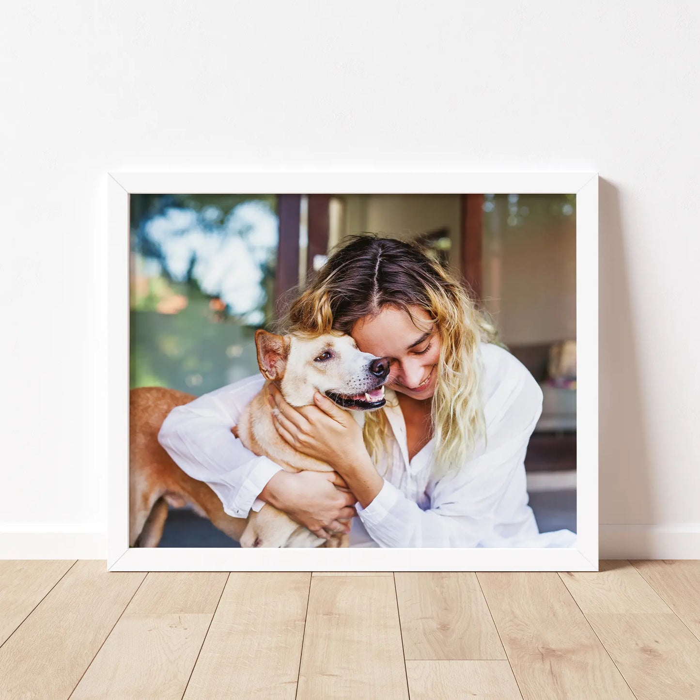 White framed horizontal photo print of a woman with wavy blonde hair hugging a small tan dog indoors, resting on a light wood floor against a neutral wall.