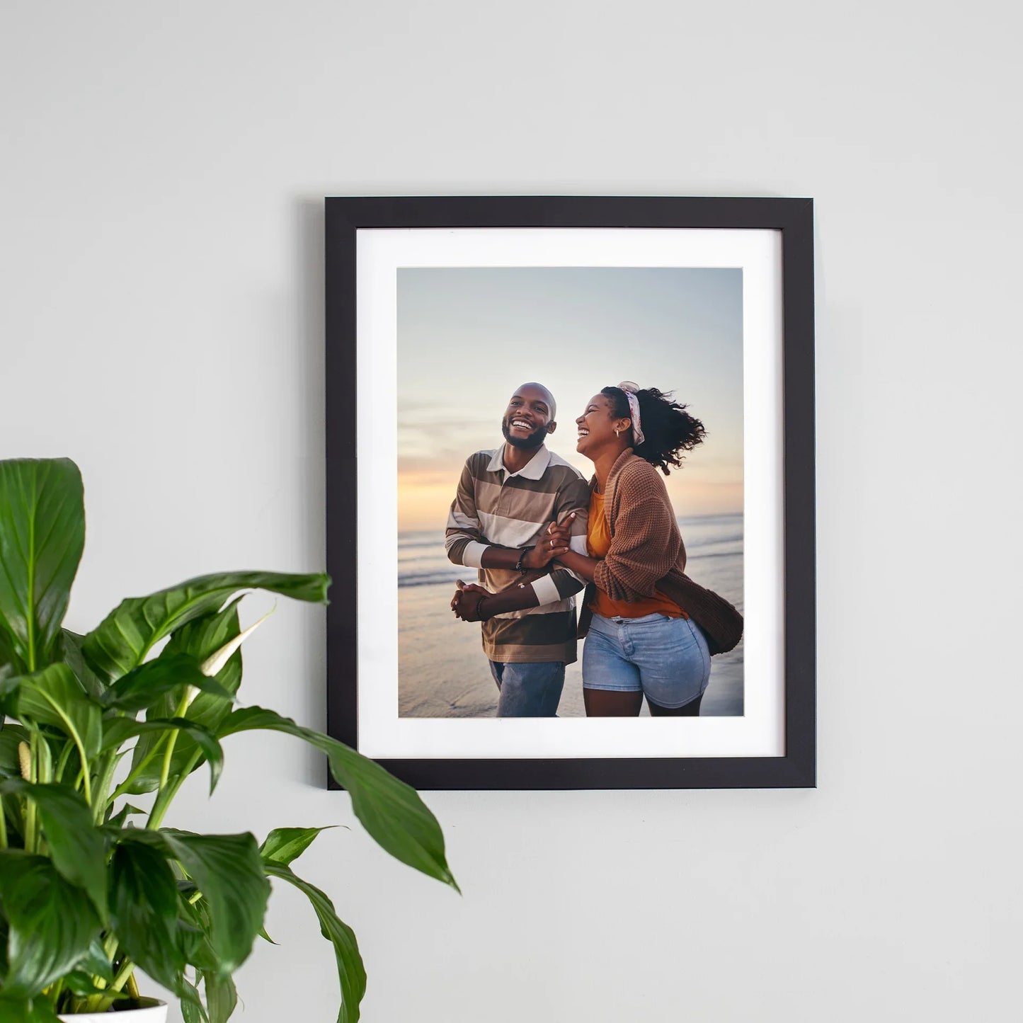 Framed print with white mat and black frame featuring couple smiling and walking on beach at sunset, vertical orientation, displayed near green houseplant.