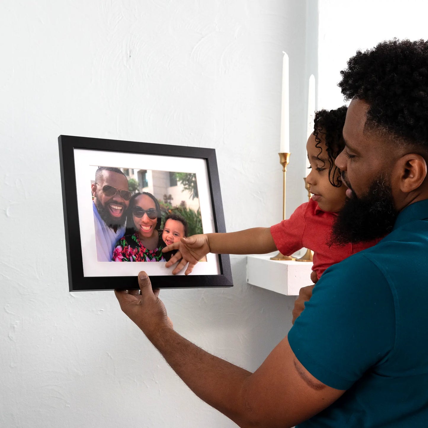 Black matted horizontal framed photo print of a smiling family portrait, held by a person while a child reaches toward the image.