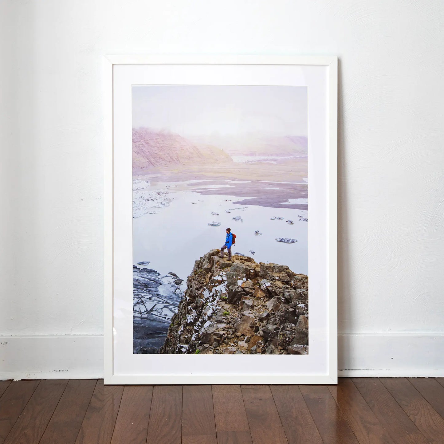White matted framed vertical photo print of a person standing on a rocky cliff above a glacier lake with misty mountains in the background.