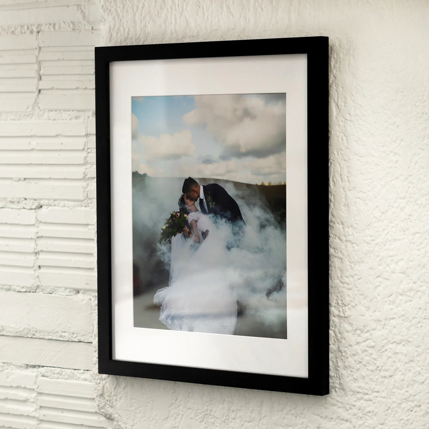 Black matted vertical framed photo of a couple embracing in wedding attire surrounded by white smoke, mounted on a textured exterior wall.