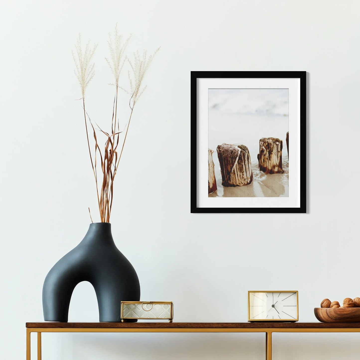 Black matted square framed photo of weathered wooden posts in shallow ocean water, displayed above a console table with decorative objects.