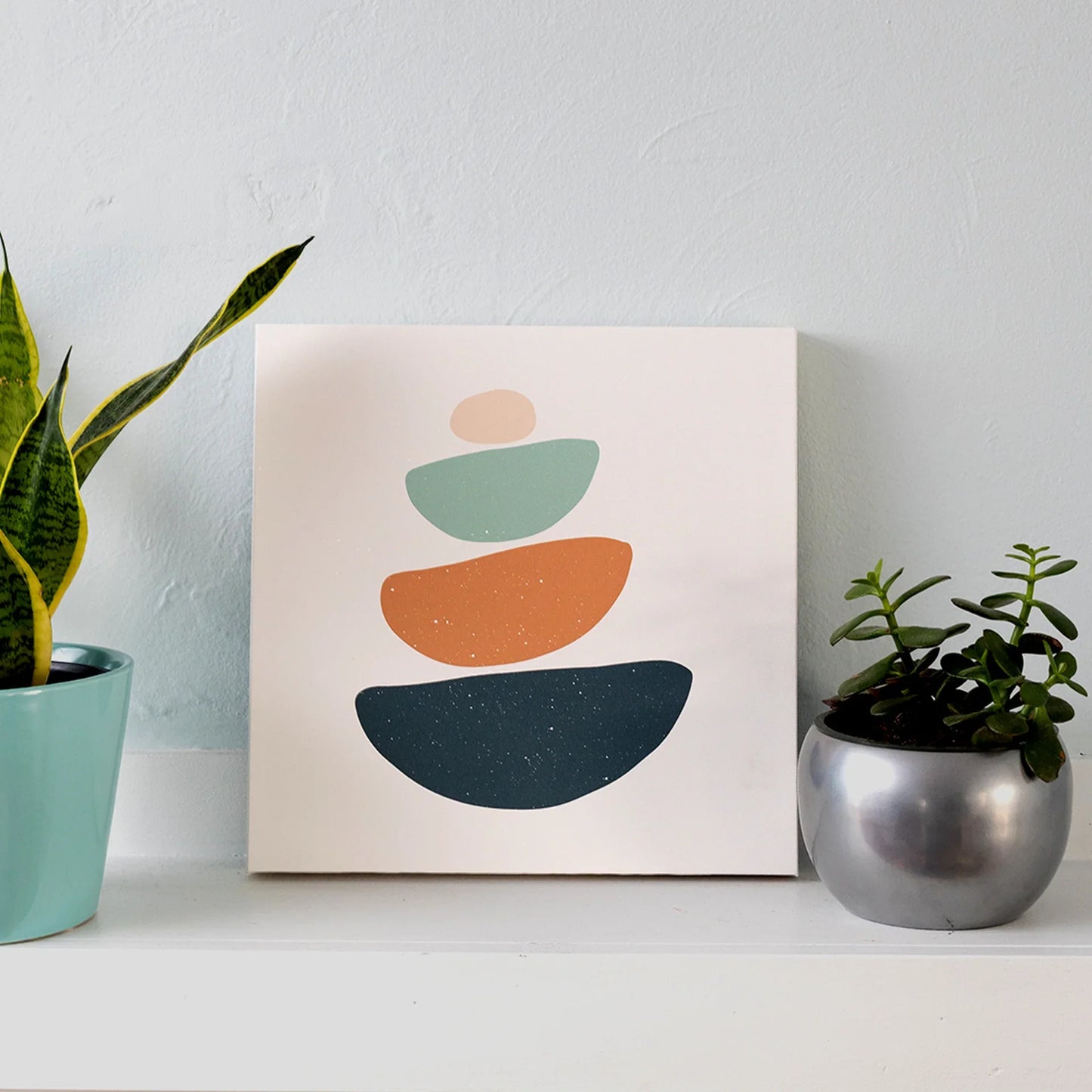 Gallery wrapped canvas featuring abstract stacked shapes in muted blue, green, and terracotta tones, displayed on a white shelf between houseplants.