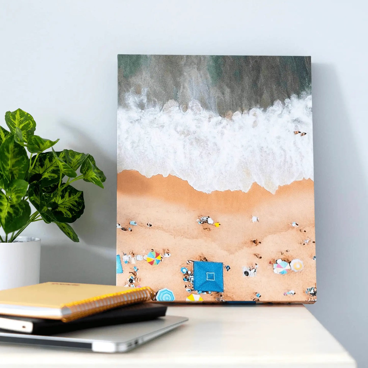 Gallery wrapped canvas print of an aerial beach scene with waves, umbrellas, and people on sand, displayed on a desk beside notebooks and a plant.