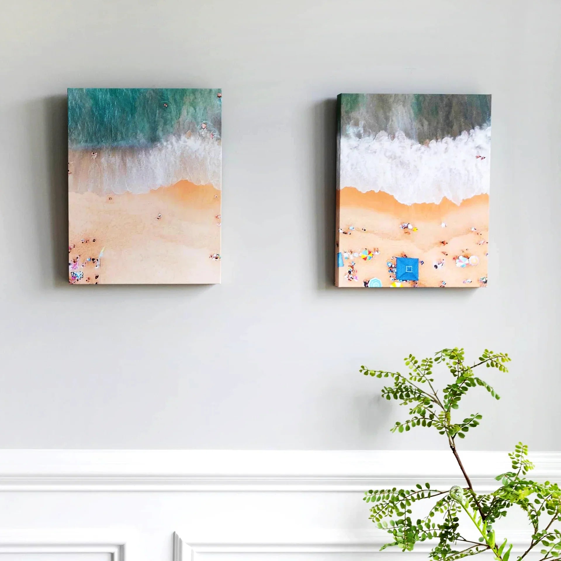 Two gallery wrapped canvas prints showing aerial beach shoreline views with waves and sand, mounted on a light gray wall above decorative molding.
