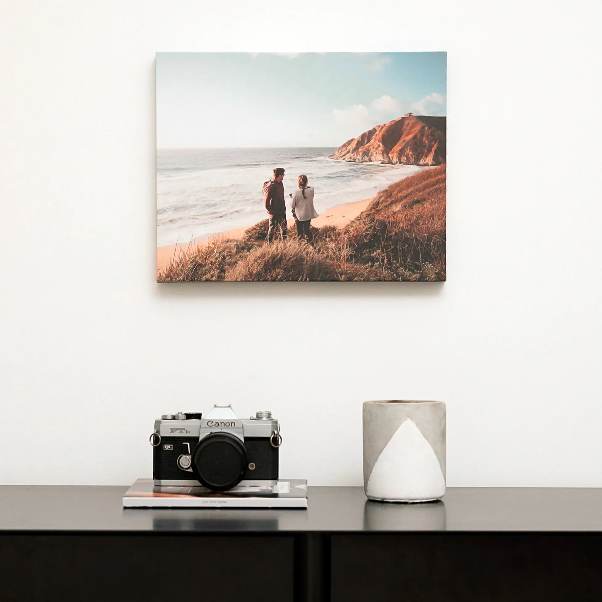 Gallery wrapped canvas featuring a couple standing on coastal cliffs overlooking the ocean, mounted above a console table with camera and ceramic decor.