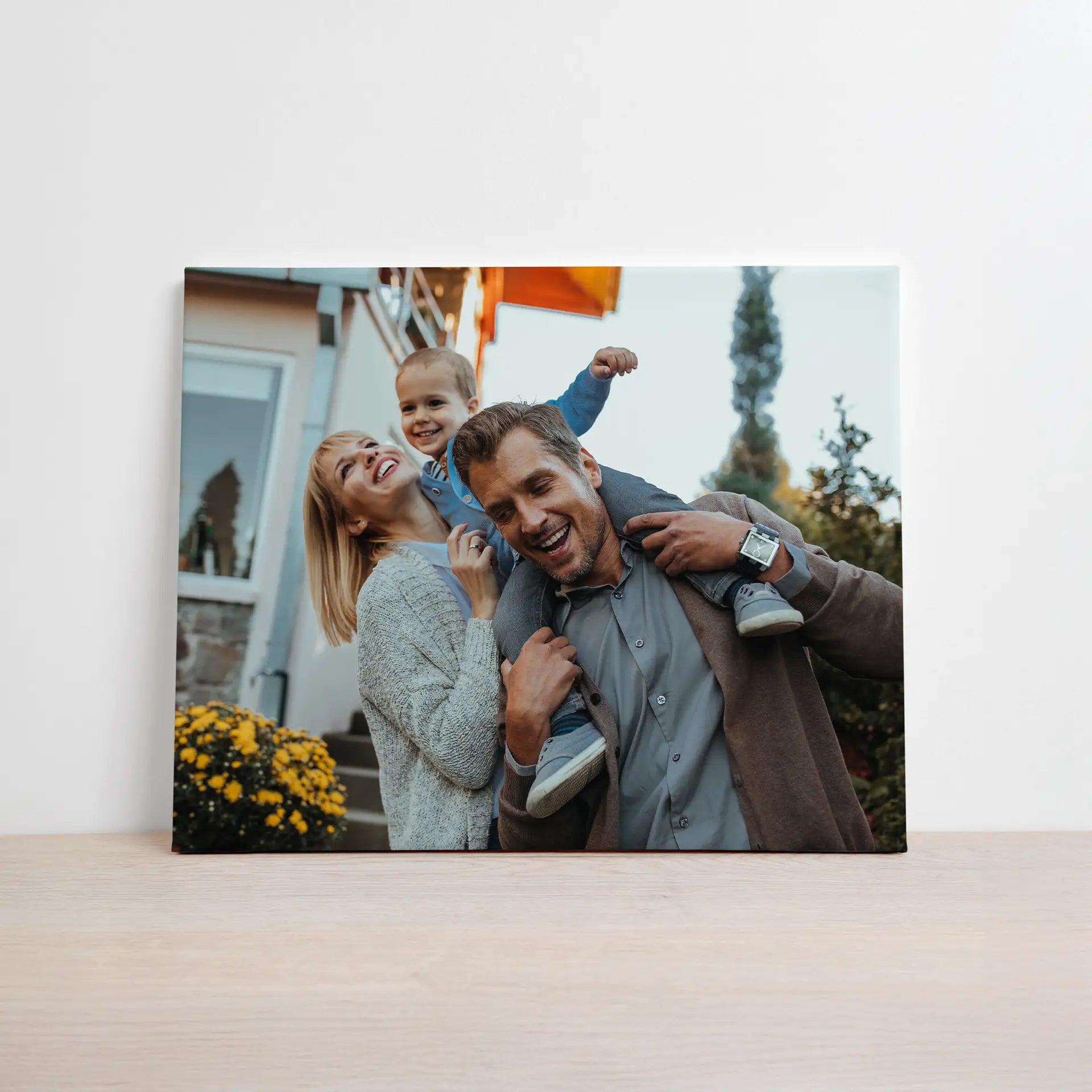 Canvas print featuring a smiling family outdoors with a child on an adult’s shoulders in front of a home, surrounded by greenery and warm light.