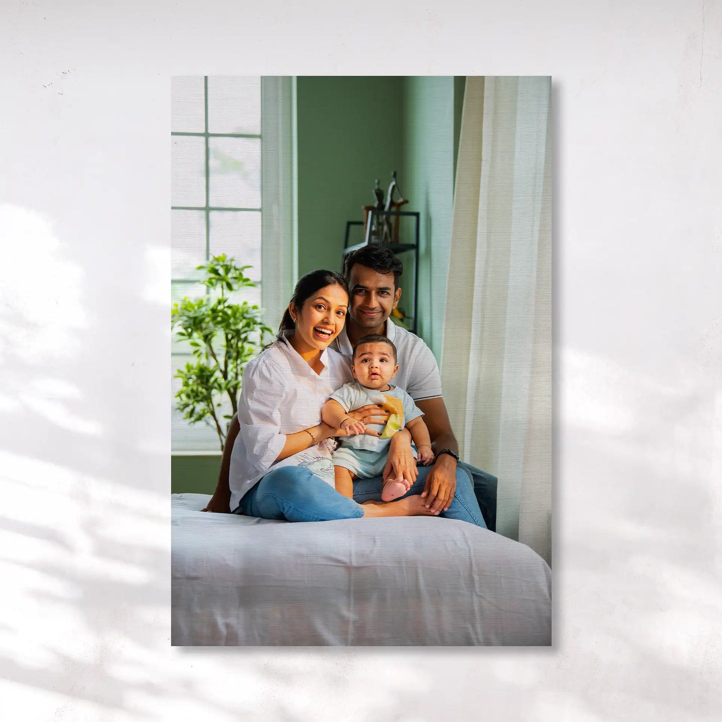 Canvas print showing a couple seated on a bed holding a baby, with soft natural light, indoor plants, and a window in the background.