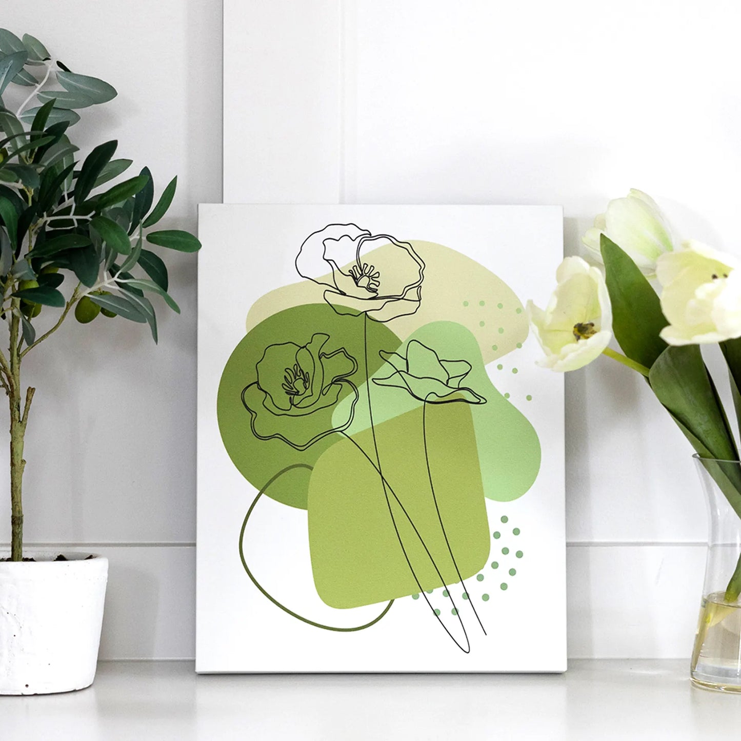 Gallery wrapped canvas with minimalist green floral line art and abstract shapes, styled on a white surface between potted plants.