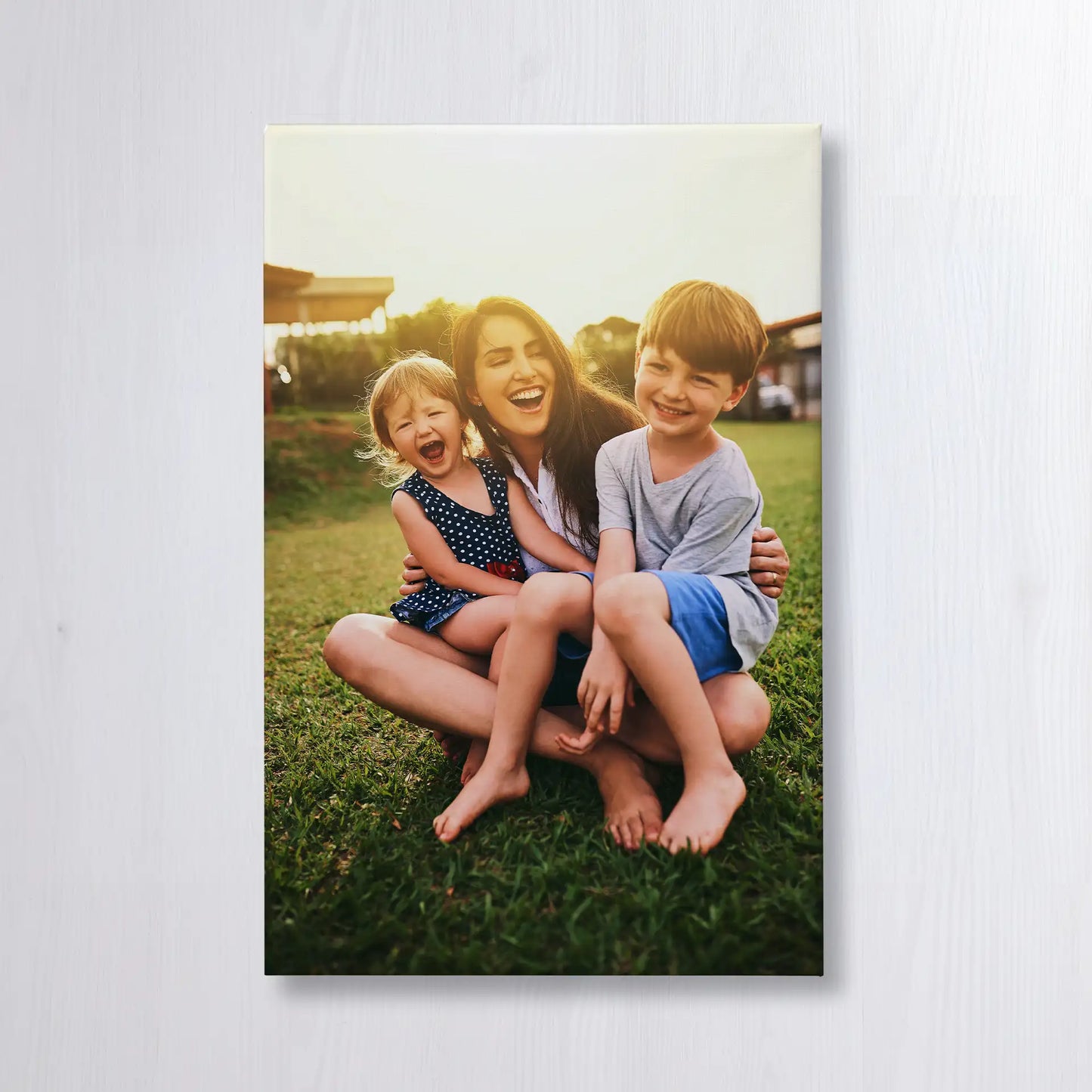 Canvas print featuring a smiling adult sitting on grass with two children, laughing together outdoors in warm sunset light.