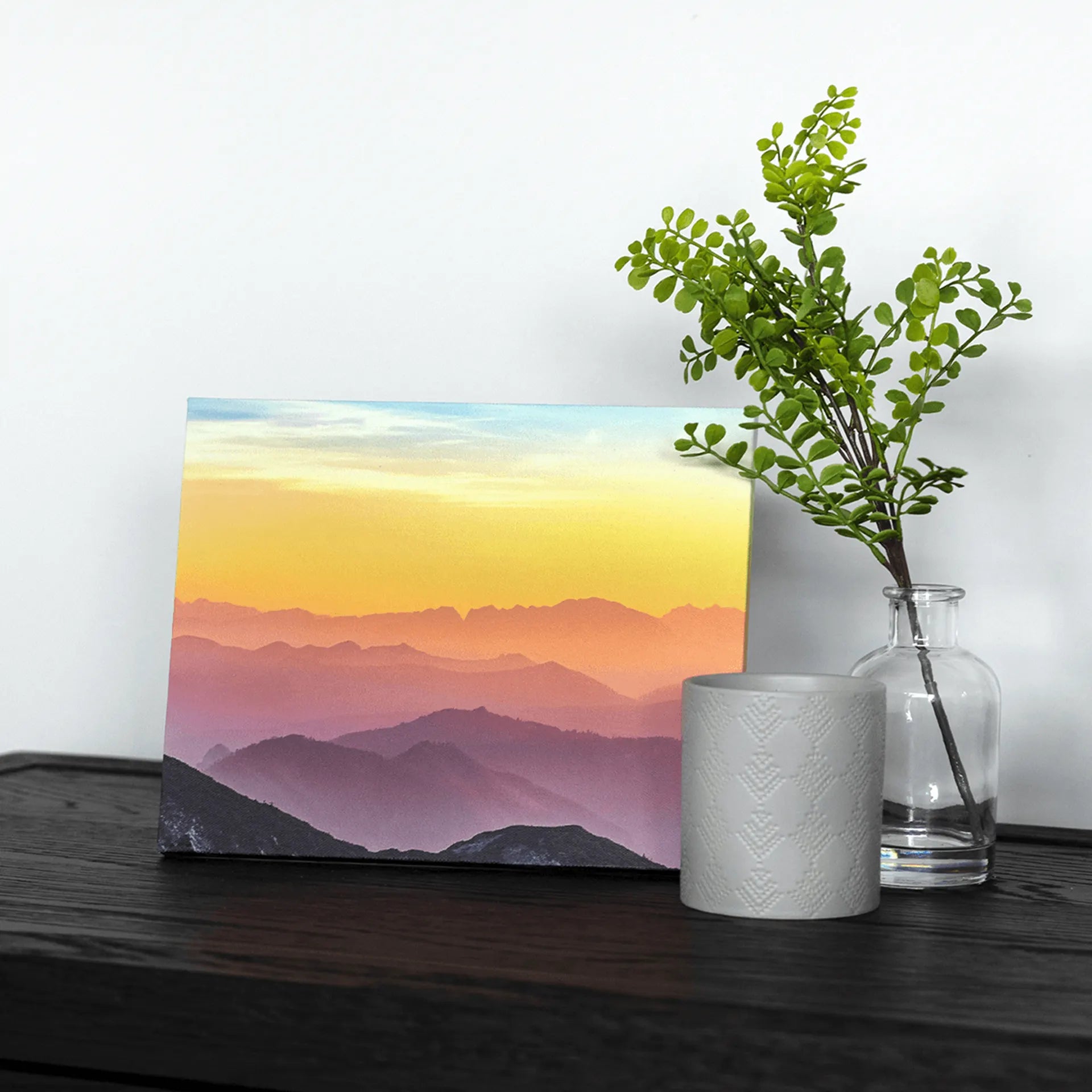 Gallery wrapped canvas print of layered mountain silhouettes at sunset in soft yellow and purple hues, styled on a dark wood surface with decorative vase.